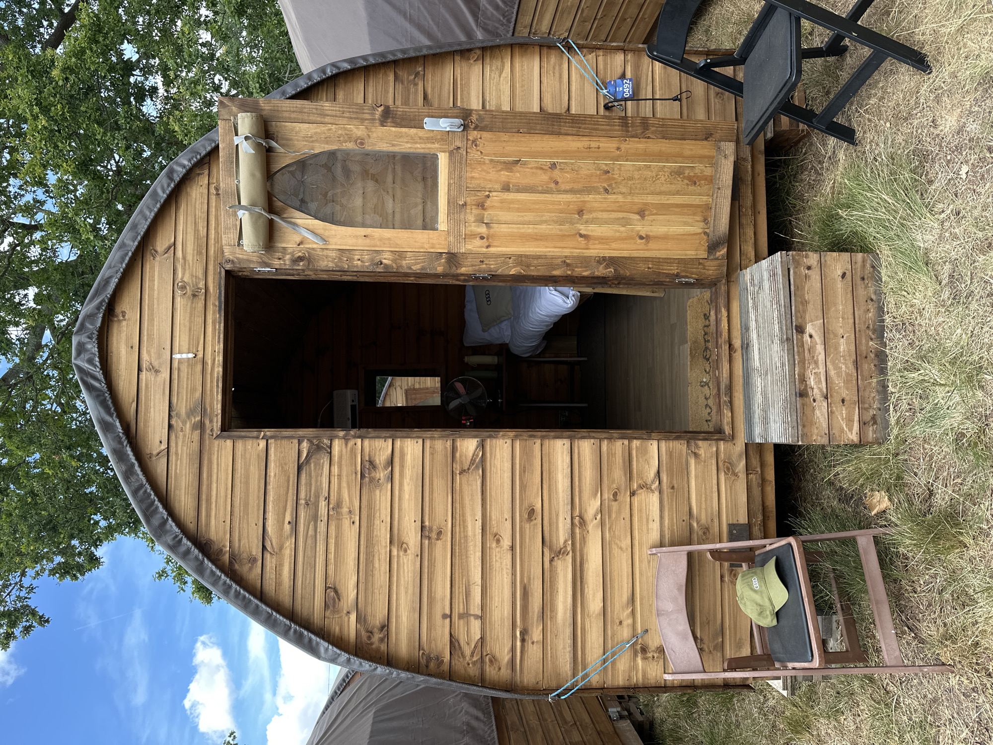 A small, wooden camping pod with a rounded roof is shown outdoors. The pod's exterior is constructed of horizontal wooden planks. A door is open, revealing a glimpse of a bed inside. To the left of the entrance, a brown folding chair with a green baseball cap on it is positioned on the grass. To the right of the entrance, a black folding chair is visible. A wooden step leads up to the entrance. The scene is set in a grassy area with trees in the background and a blue sky with clouds above. A tag with the number "0492" is attached to the side of the pod.
