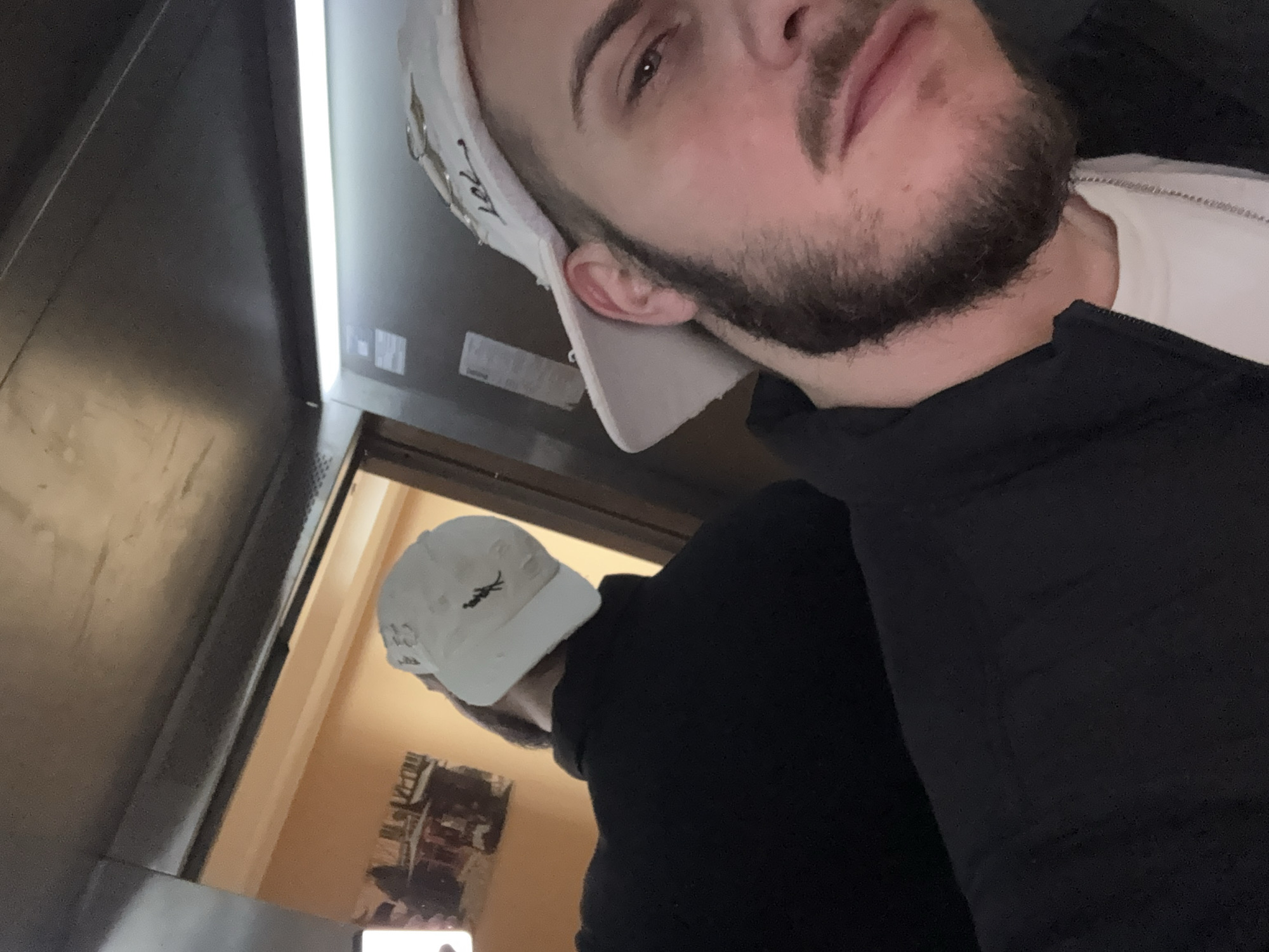 A selfie of a light-skinned male wearing a white baseball cap and a black jacket. He has a dark beard and is looking into the camera. The background is a metal elevator, with a bright light overhead. The reflection in the elevator shows another individual wearing a similar white baseball cap and a dark shirt. Behind that individual is a tan-walled room with a poster or artwork visible. The location cannot be determined to be in Sofia, Bulgaria based on the image alone.