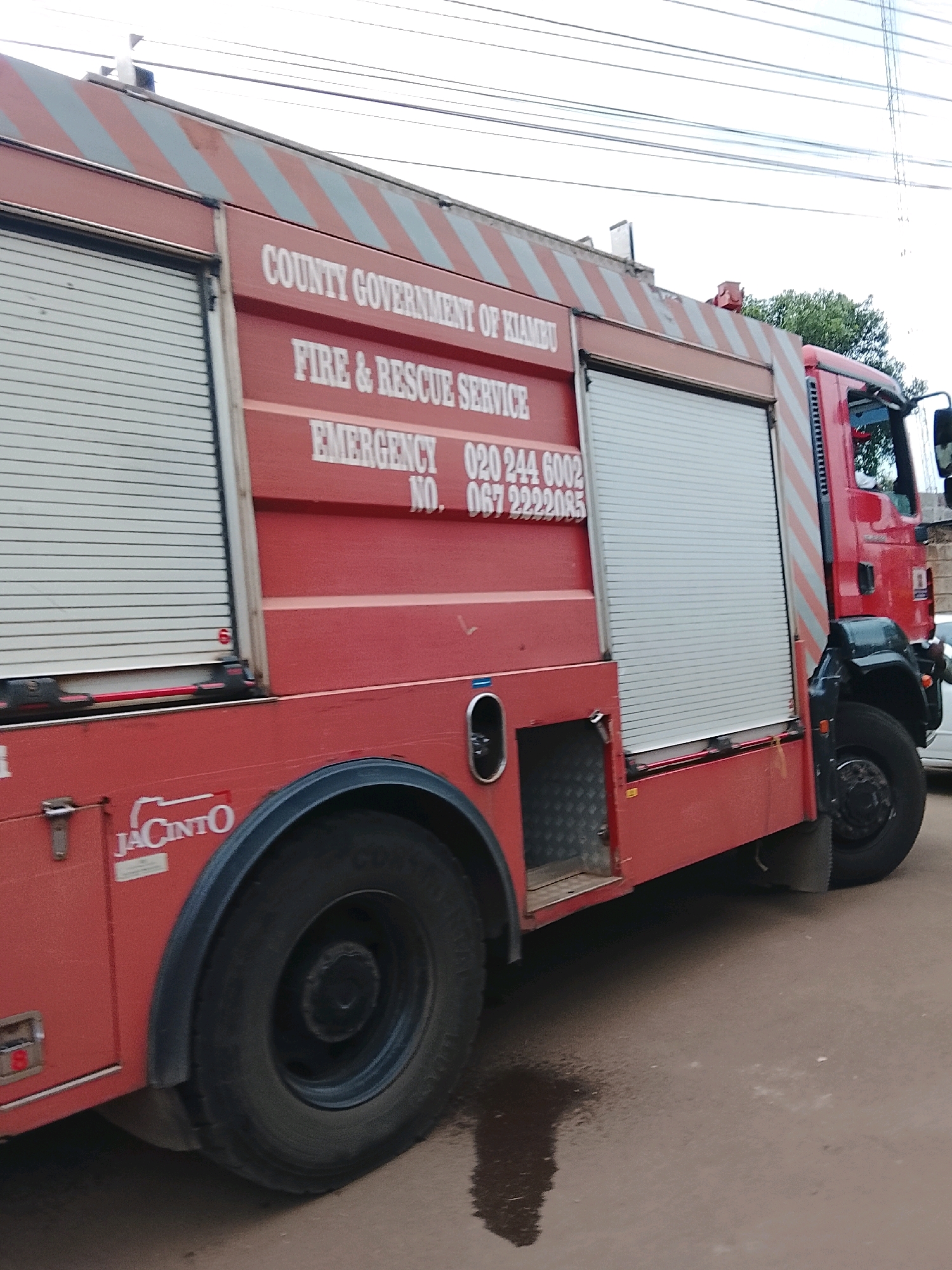 An exterior, eye-level, medium shot shows the side of a red fire truck parked on a wet, paved surface. The truck has white and gray diagonal stripes along its upper edge. Text on the side reads: "COUNTY GOVERNMENT OF KIAMBU," "FIRE & RESCUE SERVICE," and "EMERGENCY NO. 020 244 6002, 067 2222085." There are roll-up doors on the side of the truck. A logo for "JACINTO" is visible on the lower portion of the truck. A large black tire is prominently featured. In the background, power lines are visible against a bright sky, and trees and buildings are dimly lit. The overall lighting is natural and appears to be daytime.