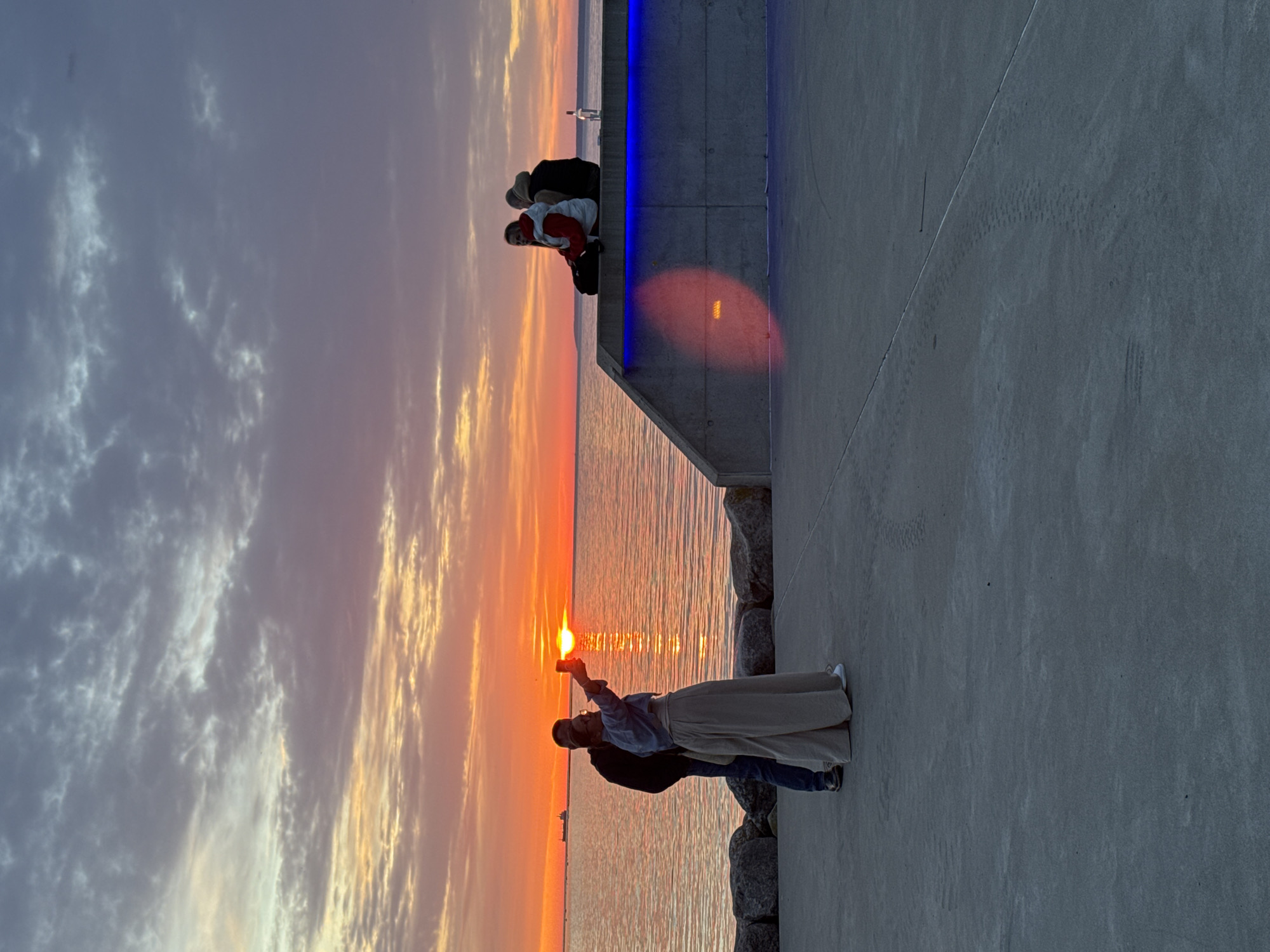 The image depicts a sunset over a body of water, likely the Baltic Sea, given the context of Tallinn, Estonia. In the foreground, a couple is taking a selfie with the sunset as a backdrop. The woman is holding up a phone to capture the photo, while the man stands beside her. They are positioned on a paved walkway near a rocky shore. Further back, two other individuals are seated on a concrete structure, observing the sunset. The structure is illuminated with blue lights. The sky displays a gradient of colors, ranging from orange and yellow near the horizon to purple and gray higher up, with scattered clouds. A ship is visible in the distance on the water. The overall scene captures a serene moment during sunset.