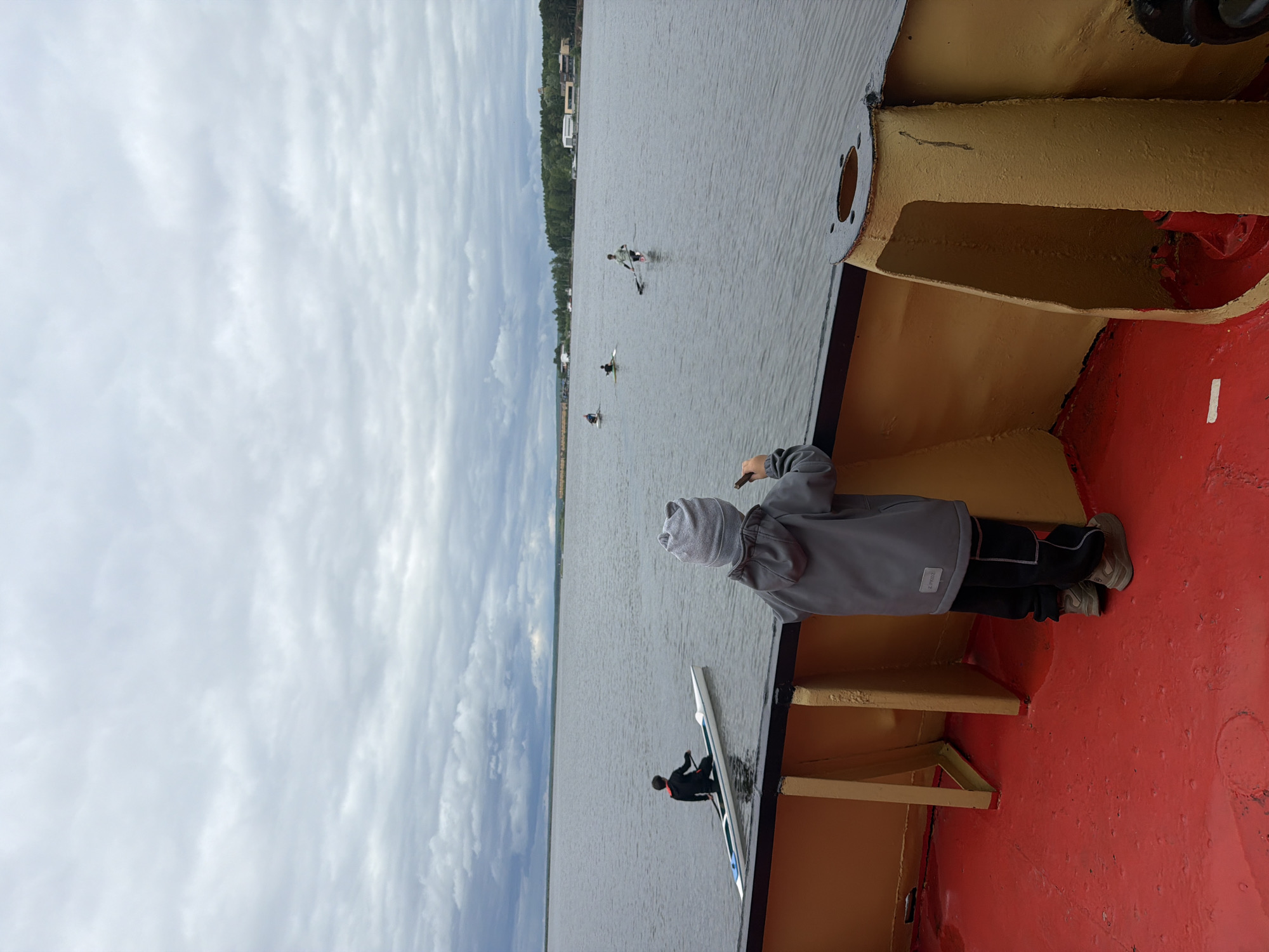 A person is standing on the deck of a boat, looking out at a body of water with several kayaks in it. The boat's deck is painted red and has some yellow supports. The person is wearing a gray jacket and hat. The sky is cloudy. The scene is likely in or near Chaikovsky, Russia, based on the location tag.