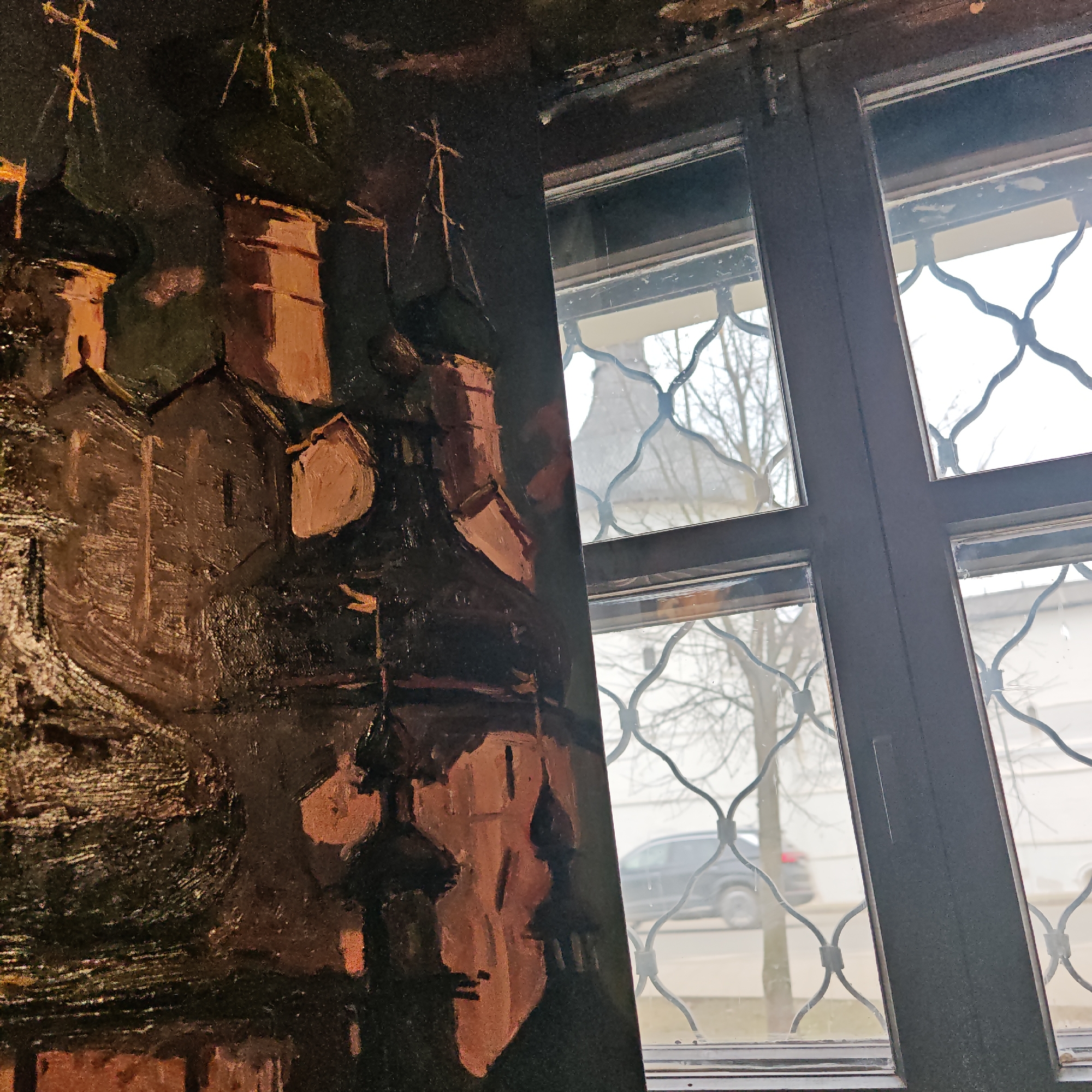The image presents a view through a multi-pane window with decorative wrought iron grillwork. Outside the window, a street scene is visible with a dark-colored car and bare trees. In the foreground, to the left of the window frame, there's a dark-toned painting depicting what appears to be a cluster of buildings with distinctive architectural features and crosses, possibly churches or religious structures. The image was taken in the city of Rostov, Russia.