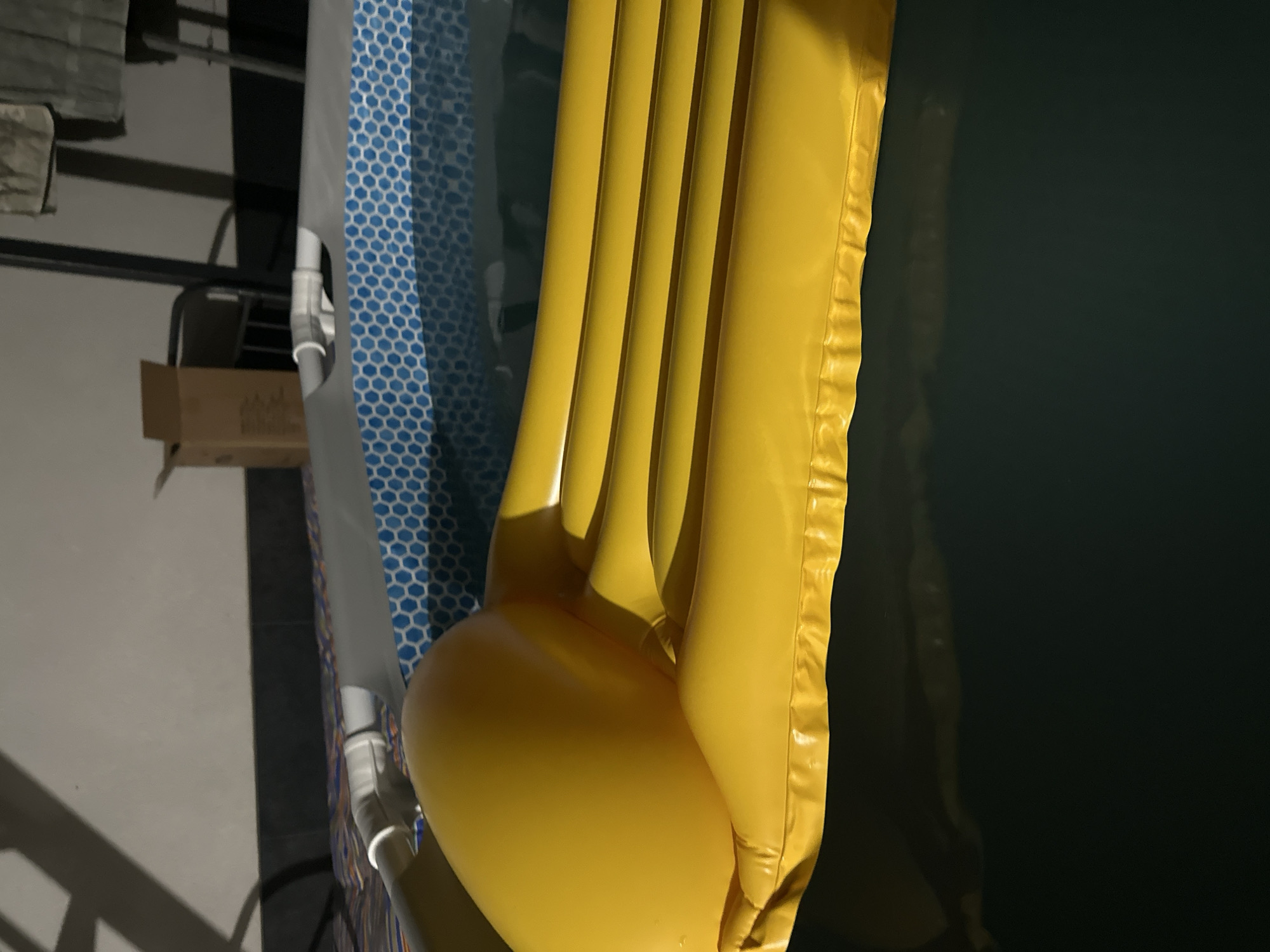 The image shows a close-up of a yellow inflatable mattress partially submerged in dark water. The mattress has a textured surface with raised ridges. To the left of the mattress, a blue and white patterned fabric is visible, possibly part of a pool liner or a covering. In the background, out of focus, are structural elements that appear to be part of an outdoor setting, including metal beams and a cardboard box. The lighting suggests it is either daytime with artificial lighting or nighttime. The scene is still and does not depict any specific activity or event. There is no visible text in the image. The context provided indicates the location as Pîrlița, Moldova.
