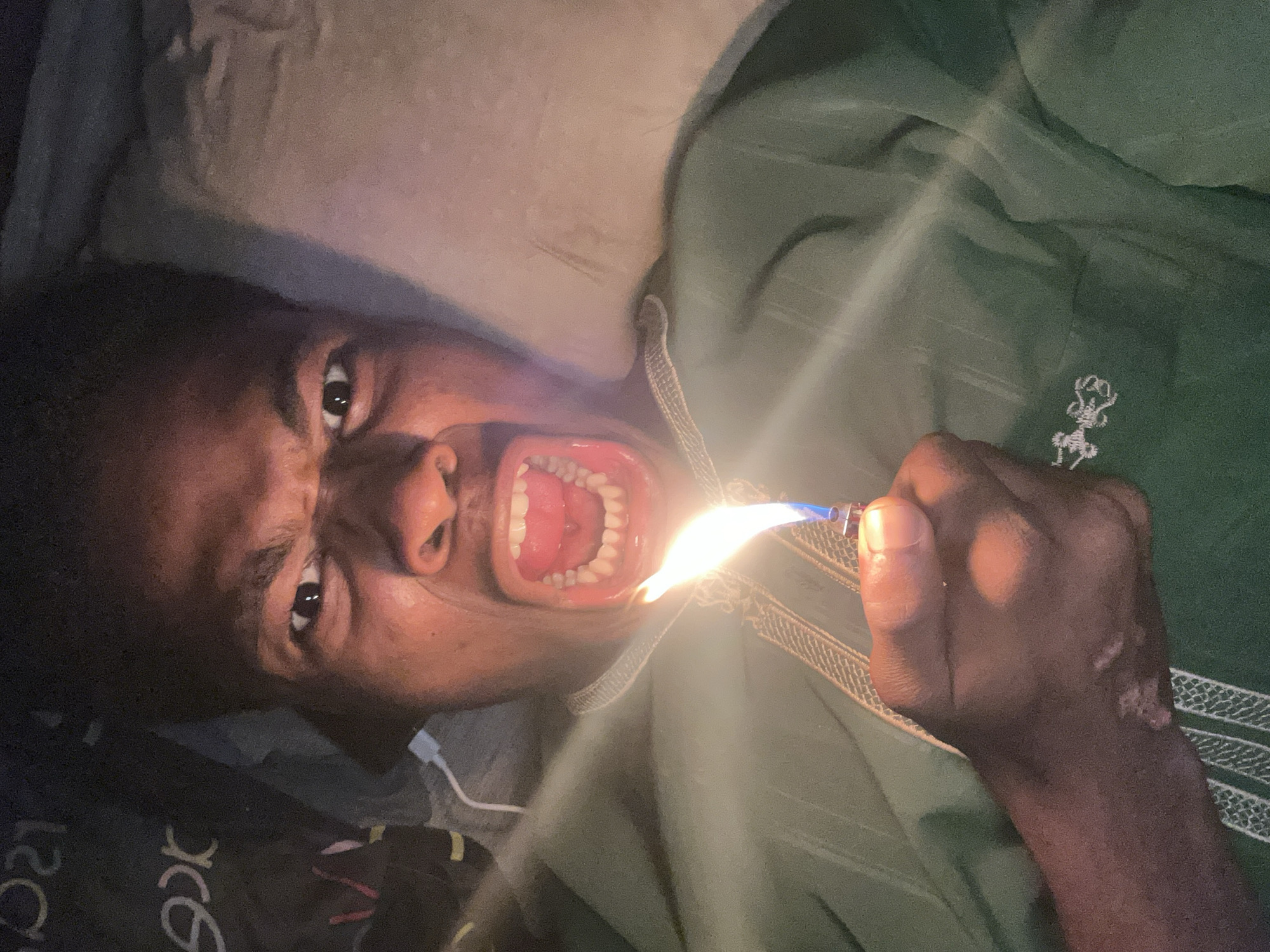 The media file depicts a close-up, low-angle shot of a person in Ihiagwa, Nigeria. The subject is lying down, possibly in bed, as indicated by a pillow or mattress in the background. They are wearing a green garment with intricate embroidery.
The person's mouth is wide open, revealing their teeth and tongue. In their right hand, they are holding a lighter, and a flame is emanating from it, directed towards their open mouth. The face shows an intense expression, with wide eyes and an open mouth that could be interpreted as surprise, pain, or a contorted expression of some kind.
The lighting suggests an indoor setting, potentially at night or in a dimly lit room, with the bright flame of the lighter being a primary light source. There are no clear indicators of weather conditions.
Visible text includes what appears to be part of a logo or writing on the person's clothing or a blanket in the foreground, but it is partially obscured and illegible. The overall impression is one of an unusual and potentially dangerous activity.
