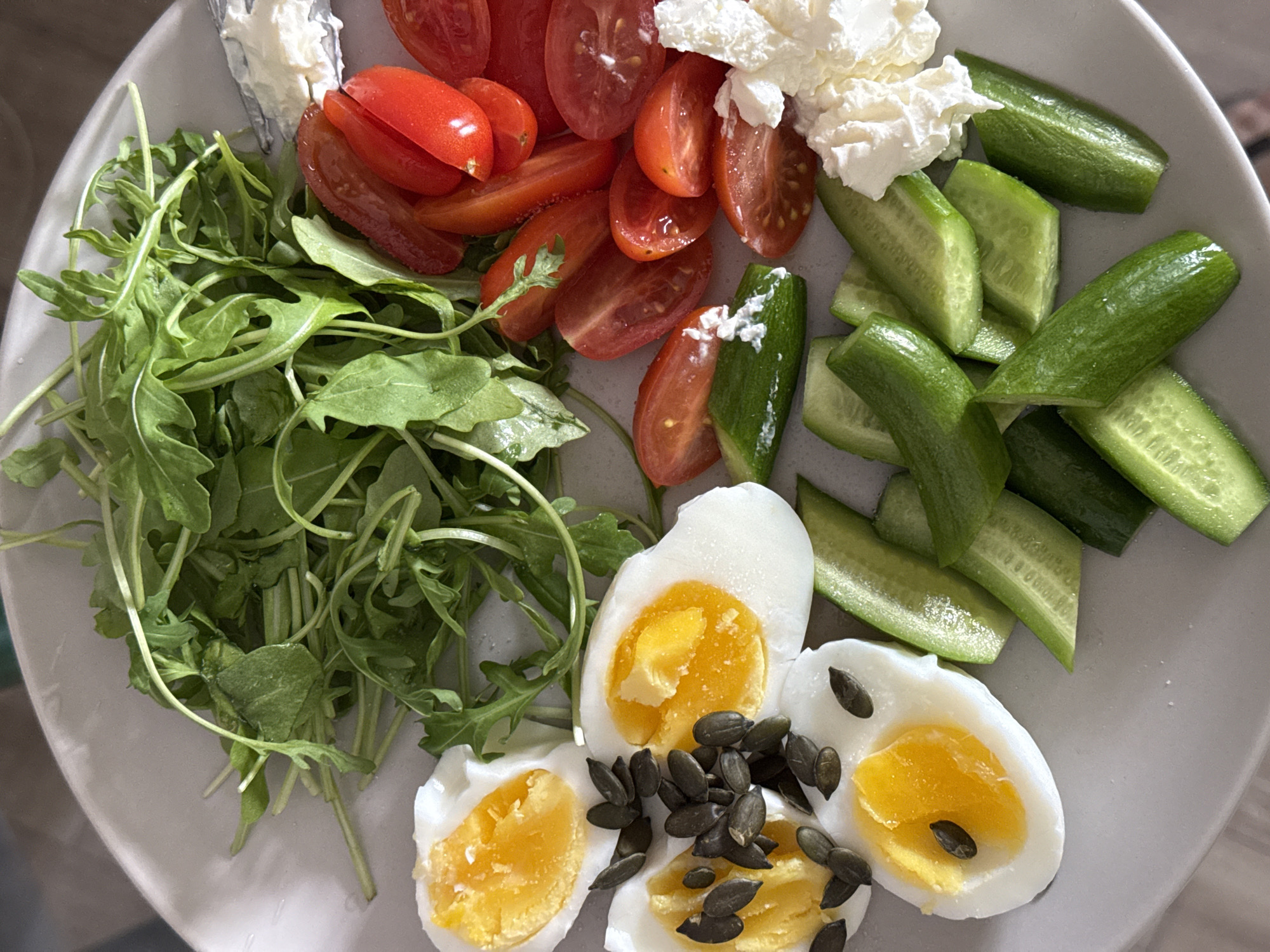 A top-down shot shows a plate of food on a table. The plate contains a pile of arugula, halved cherry tomatoes, a dollop of soft white cheese (possibly ricotta or labneh), sliced cucumbers, and three halved hard-boiled eggs topped with dark seeds (likely pumpkin seeds). The food is arranged in distinct sections on the plate. The plate appears to be gray and rests on a surface with a slightly reflective quality. The context of Tel-Aviv, Israel is not visually evident.