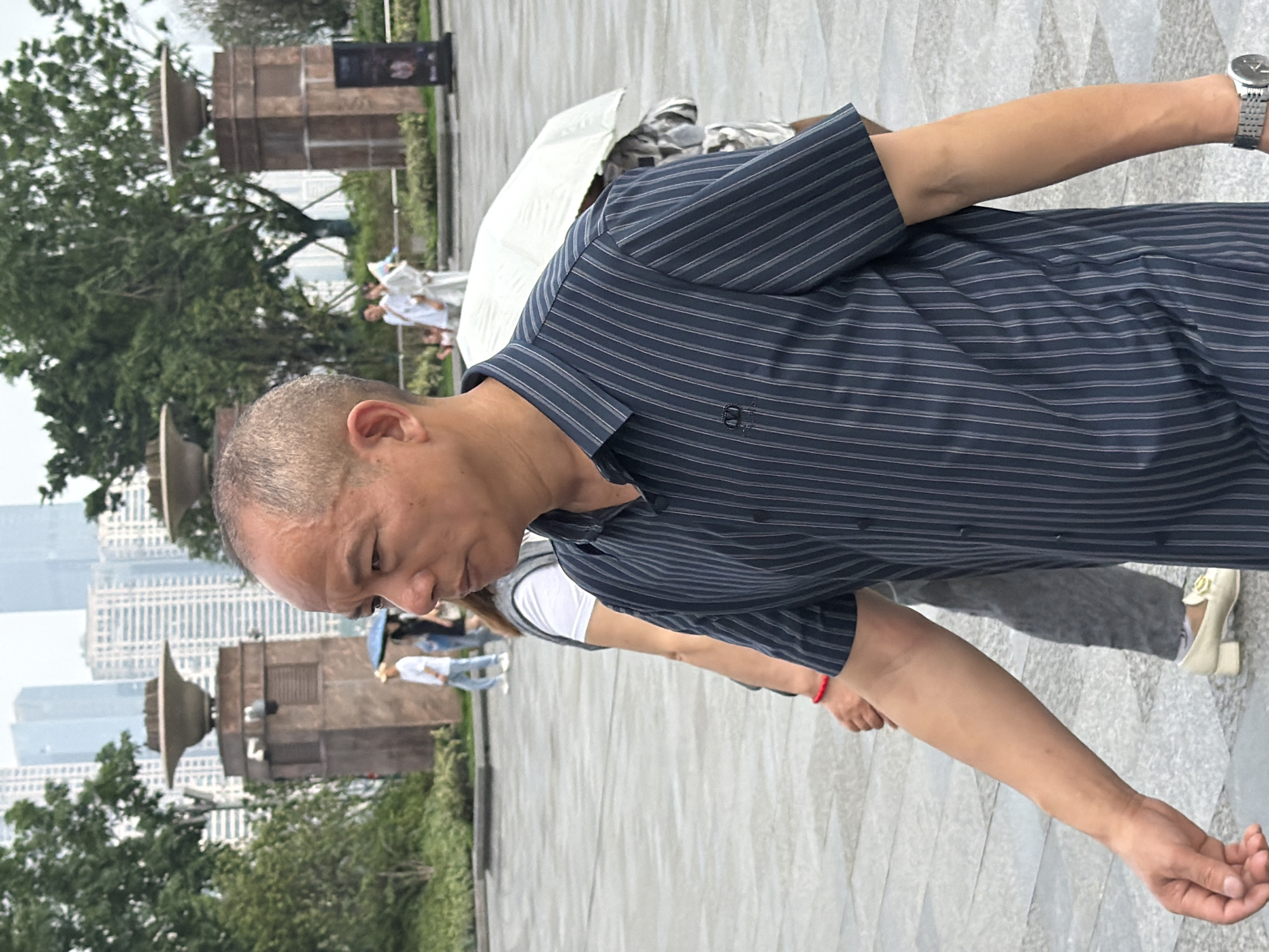 A middle-aged East Asian man, bald with short-cropped hair and visible veins on his forehead, is walking outdoors in what appears to be a city plaza or park in Yuzhong District, China. He is wearing a dark blue, short-sleeved collared shirt with thin light grey vertical stripes and a subtle embroidered logo on the left chest. His right arm is extended slightly forward.
Behind him, a person is partially visible, wearing a white t-shirt and grey pants, with a red bracelet on their left wrist. Further in the background, other individuals are seen walking, some with umbrellas, suggesting overcast or rainy weather. The environment includes large planters with decorative tops, trees, and modern high-rise buildings in the distance, characteristic of an urban landscape. The ground is paved with grey tiles.
