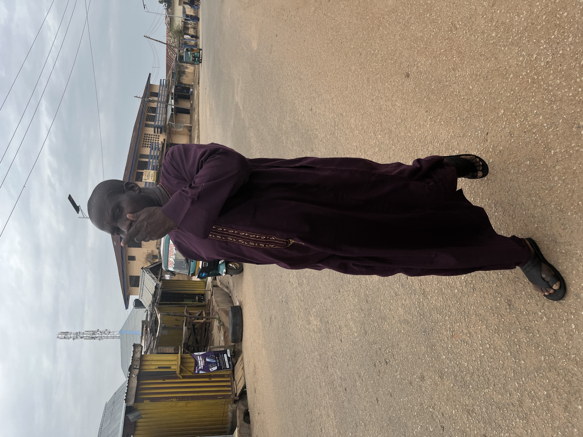 An individual wearing a purple robe and sandals is standing on an asphalt road in what appears to be a commercial area of Jos, Nigeria. The person is obscuring their face with their hand. Buildings of various heights and architectural styles line the road, including structures with corrugated metal roofs and more substantial multi-story buildings. A communications tower is visible in the background. A "Keke Napep," or tricycle transport, is present in the frame.