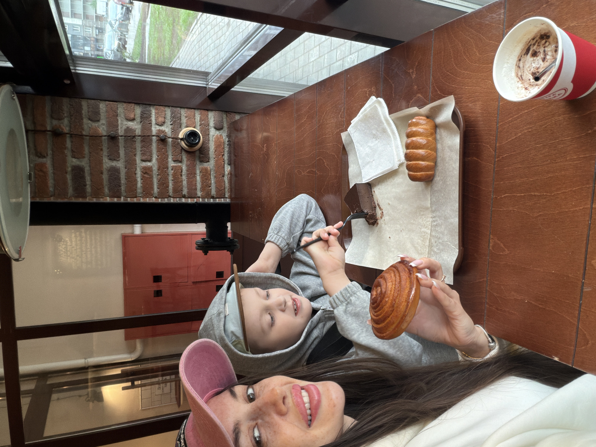 The image shows a woman in a pink baseball cap and a boy in a gray hooded sweatshirt taking a selfie indoors. The boy is eating chocolate cake with a fork, and the woman is holding a cinnamon roll. A tray with a pastry and a napkin, and a red and white cup of coffee are on the brown wooden table. The room has a brick wall and a window offering an exterior view. The location is Saint Petersburg, Russia.