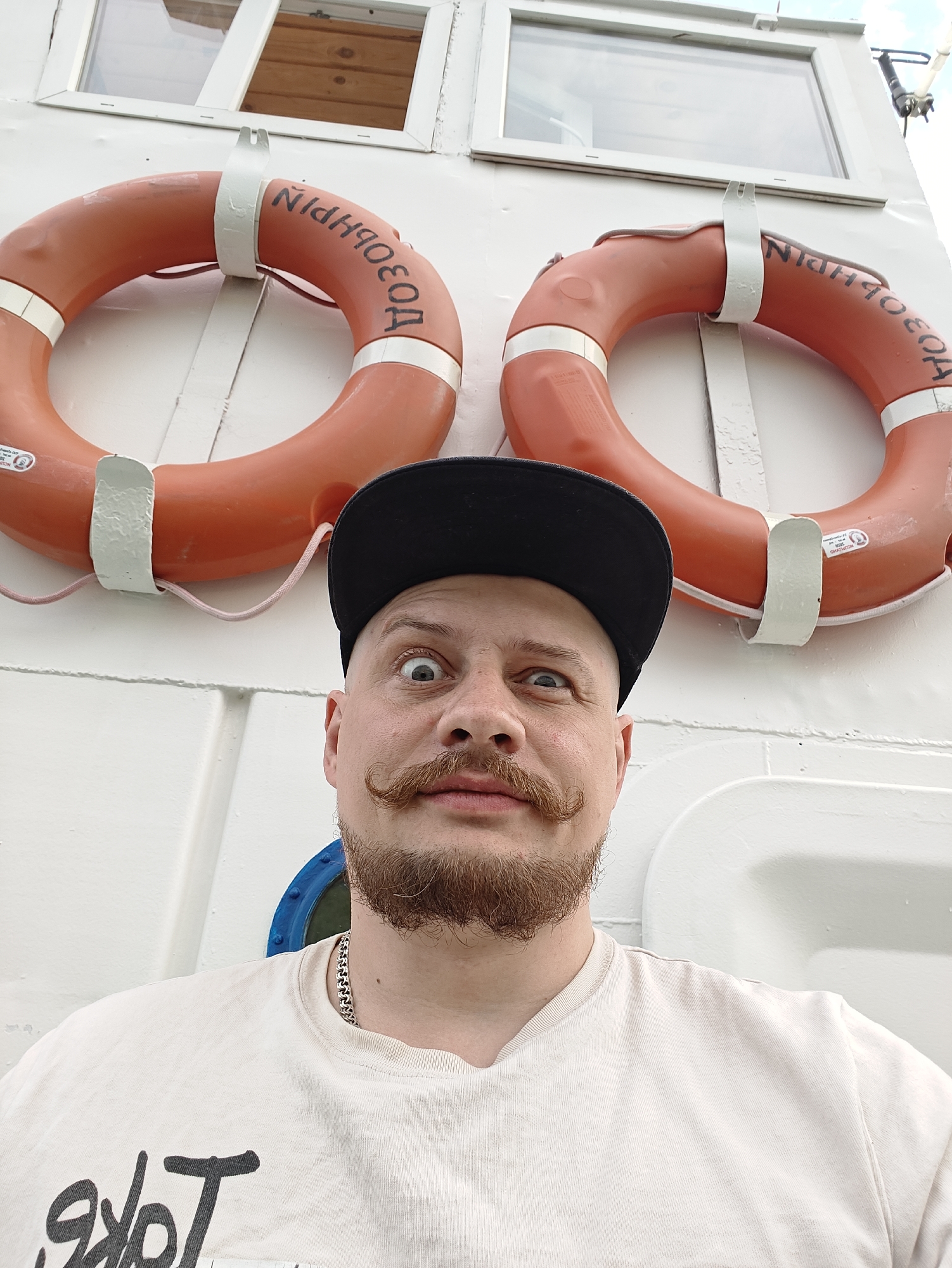 A man with a handlebar mustache and a black baseball cap is taking a selfie in front of a white wall with two orange life preservers. The life preservers have Cyrillic lettering on them, which appears to read "НОЗОБОР." There is a window above the life preservers, and a blue-rimmed circular window visible to the left of the man's face. The man is wearing a light-colored t-shirt with a black graphic design and a silver chain necklace. The image appears to be taken on a boat or a pier, but no specific location in Manat, Kazakhstan can be determined from the visual evidence.