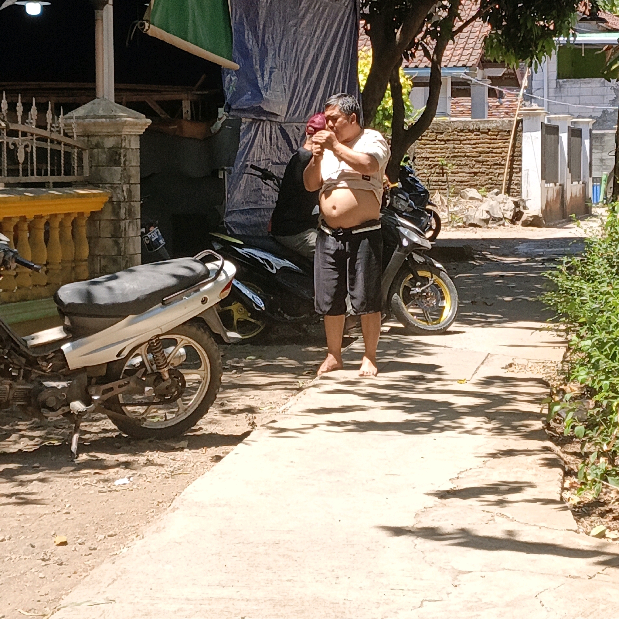 The image depicts an outdoor scene in Mangli, Indonesia, under bright sunlight. In the foreground, a light-colored motorcycle with a black seat is parked on a dirt path. To the right of the motorcycle, a man stands barefoot on a concrete walkway. He is wearing a light-colored t-shirt that is lifted up, exposing his abdomen, and dark shorts. The man is holding what appears to be a cigarette and is in the process of lighting it. Behind the man, another motorcycle, mostly black with yellow accents on its wheels, is visible. A dark blue tarp is draped over a structure behind the motorcycles. The background shows a brick wall, a tiled roof of a building, and trees with dappled sunlight casting shadows on the ground. A decorative yellow fence is present on the left side of the image. The overall environment suggests a residential or semi-commercial area.