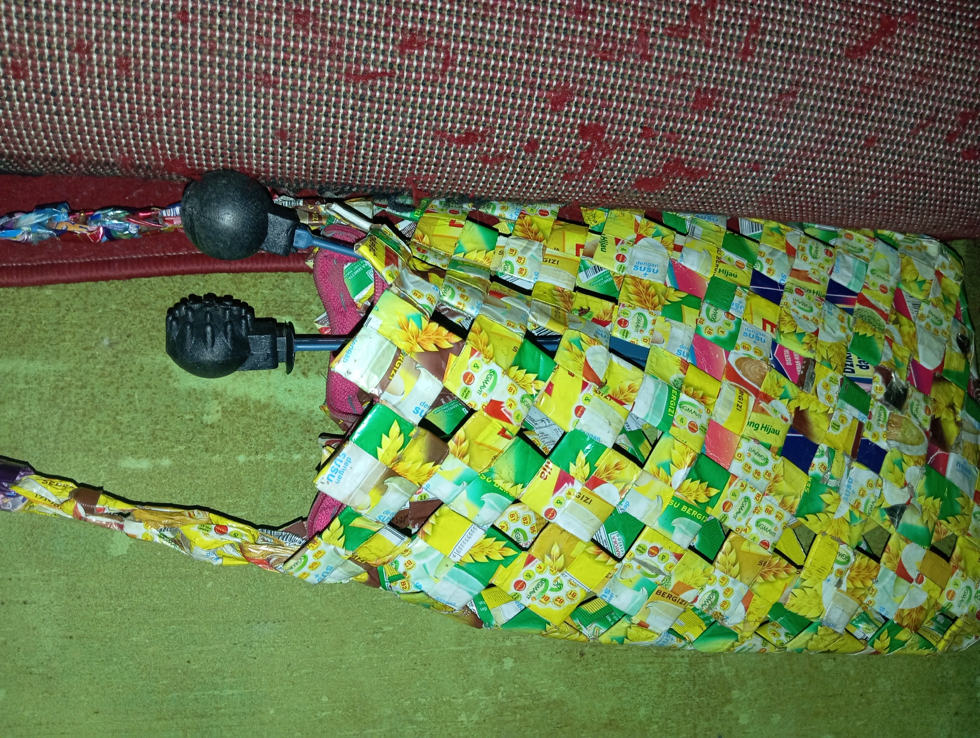 The image shows a close-up view of a handmade bag crafted from recycled wrappers, likely plastic packaging from snacks or food items. The bag is woven with strips of brightly colored wrappers, predominantly yellow, green, and white, creating a patchwork effect. The bag has a strap also made from the same recycled materials, and it appears to be hanging from something. Two black, spherical objects, possibly umbrella handles, are visible behind the bag, partially obscuring it. The background consists of a textured, green wall on the left and a red and black patterned fabric on the right. The lighting is dim and appears to be artificial. There are no people or animals visible in the image, and no specific location in Rejosari, Indonesia can be identified from the visual information.