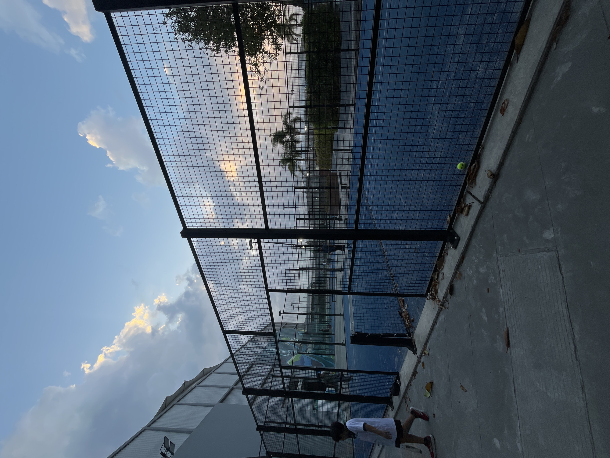 The photograph presents an outdoor padel tennis court in Unknown, Unknown. The court is enclosed by a black wire mesh fence. A person is visible near the bottom right corner of the frame, wearing a white shirt and dark shorts with red shoes. The concrete ground is littered with fallen leaves. Through the fence, another court is faintly visible in the background. The sky is partly cloudy, with the sun casting a glow on the clouds.