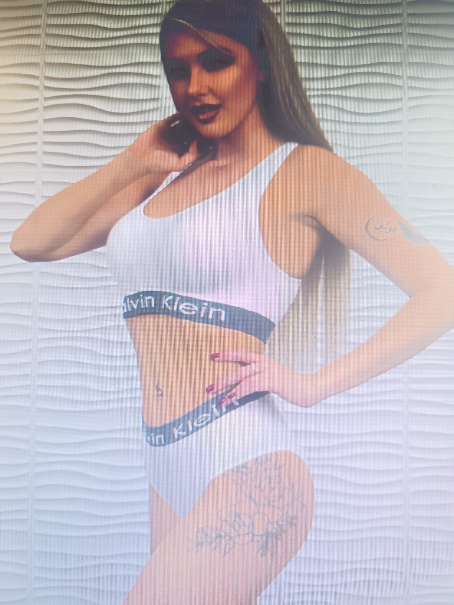 A woman is pictured in Klyuchi, Russia, posing provocatively against a textured white wall. She is wearing white Calvin Klein underwear, consisting of a sports bra and bikini briefs, both with the brand's signature grey waistband. Her right hand is on her neck, while her left hand rests on her hip. A small tattoo is visible on her left arm, and a floral tattoo adorns her right thigh. Her makeup is dramatic, with dark lipstick and eye shadow. The lighting is soft, creating a diffused effect.