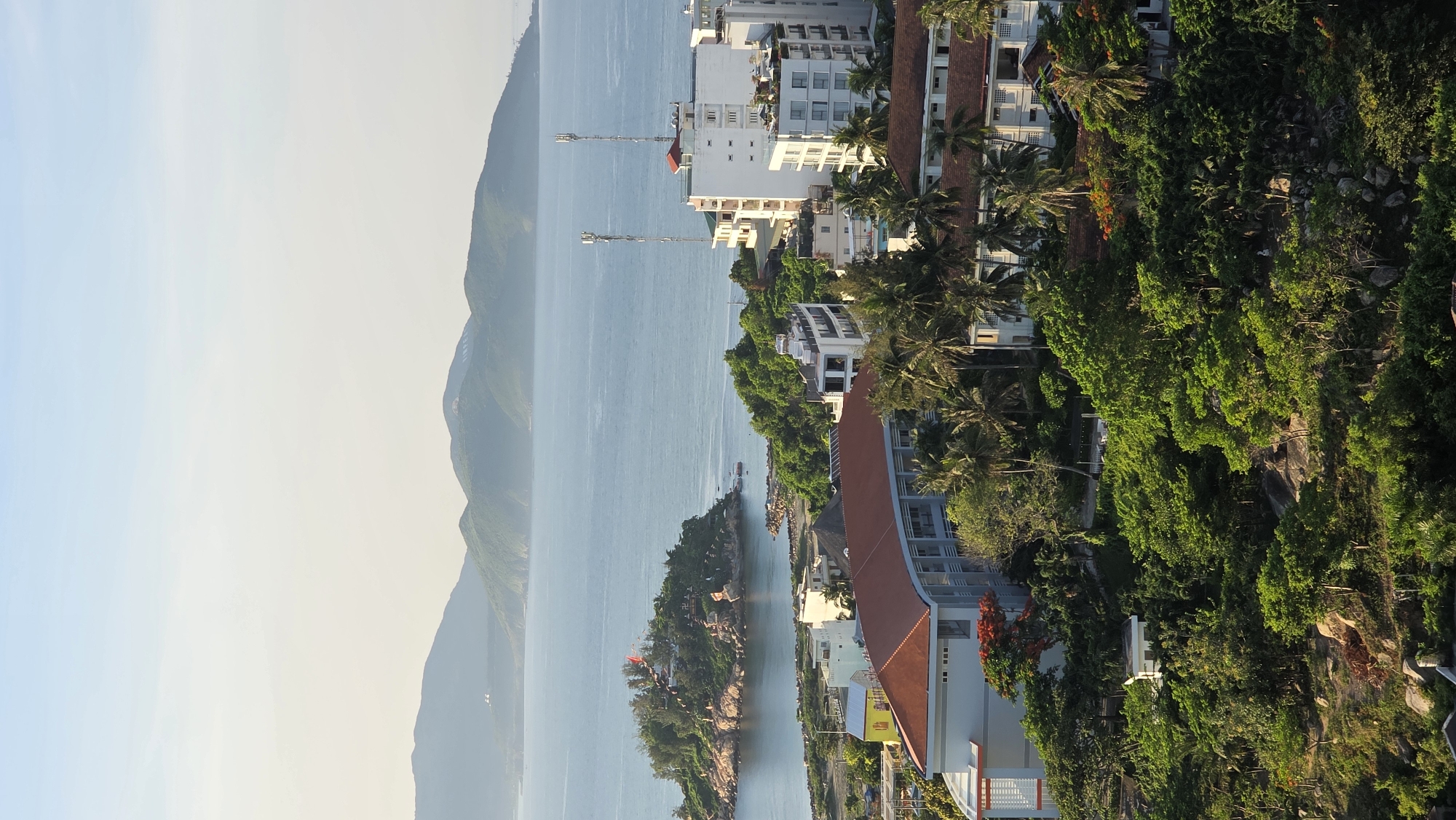 The image depicts a panoramic view of a coastal city, likely Nha Trang, Vietnam, taken from a high vantage point. The scene is divided into distinct layers: the sky, the sea, a small island, and the cityscape.
The sky is a soft, gradient of pale blue and white, suggesting either early morning or late afternoon light. Below the sky, a series of undulating, green mountains form the distant horizon, their peaks softened by atmospheric haze.
The central focus is the calm, blue sea, which stretches across the middle ground. On the left side of the sea, a small, densely vegetated island rises prominently. This island is adorned with numerous small structures, a red flag flying from a pole at its highest point, and what appear to be stone pathways and decorative elements. The shoreline of the island is rocky, with some areas of exposed sand.
In the foreground and mid-ground, the cityscape is visible. Modern multi-story buildings, predominantly white and light-colored, are clustered together. Some buildings feature balconies and numerous windows. One particularly prominent building in the mid-ground has a distinctive curved structure with a red-tiled roof and light blue walls. Adjacent to these buildings are areas with lush green trees, including palm trees, and patches of red flowers, indicating a tropical or subtropical climate. Two tall cellular towers are visible, standing out against the skyline on top of one of the white buildings. The lower part of the image shows a hillside covered in dense green foliage and large rocks.
The overall impression is one of a coastal urban environment integrated with natural elements, bathed in soft, natural light. There are no visible people or specific human activities captured in the frame. The location is identified as Bac Nha Trang Ward, Vietnam, which is a coastal district in the city of Nha Trang, known for its beaches and scenic beauty.