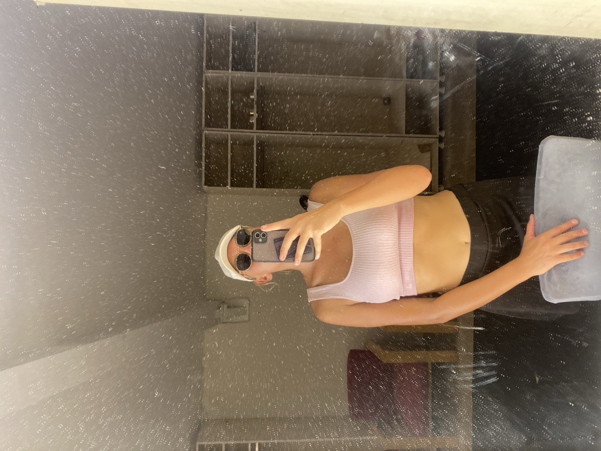 A young woman wearing a white baseball cap, sunglasses, and a pink crop top stands in front of a mirror, holding up a smartphone to take a selfie. She is also wearing a black skirt. The mirror is dirty, with numerous water spots and smudges. In the background, visible in the mirror's reflection, are lockers, a chair, and what appears to be a changing room or gym area. The overall environment suggests a fitness facility.