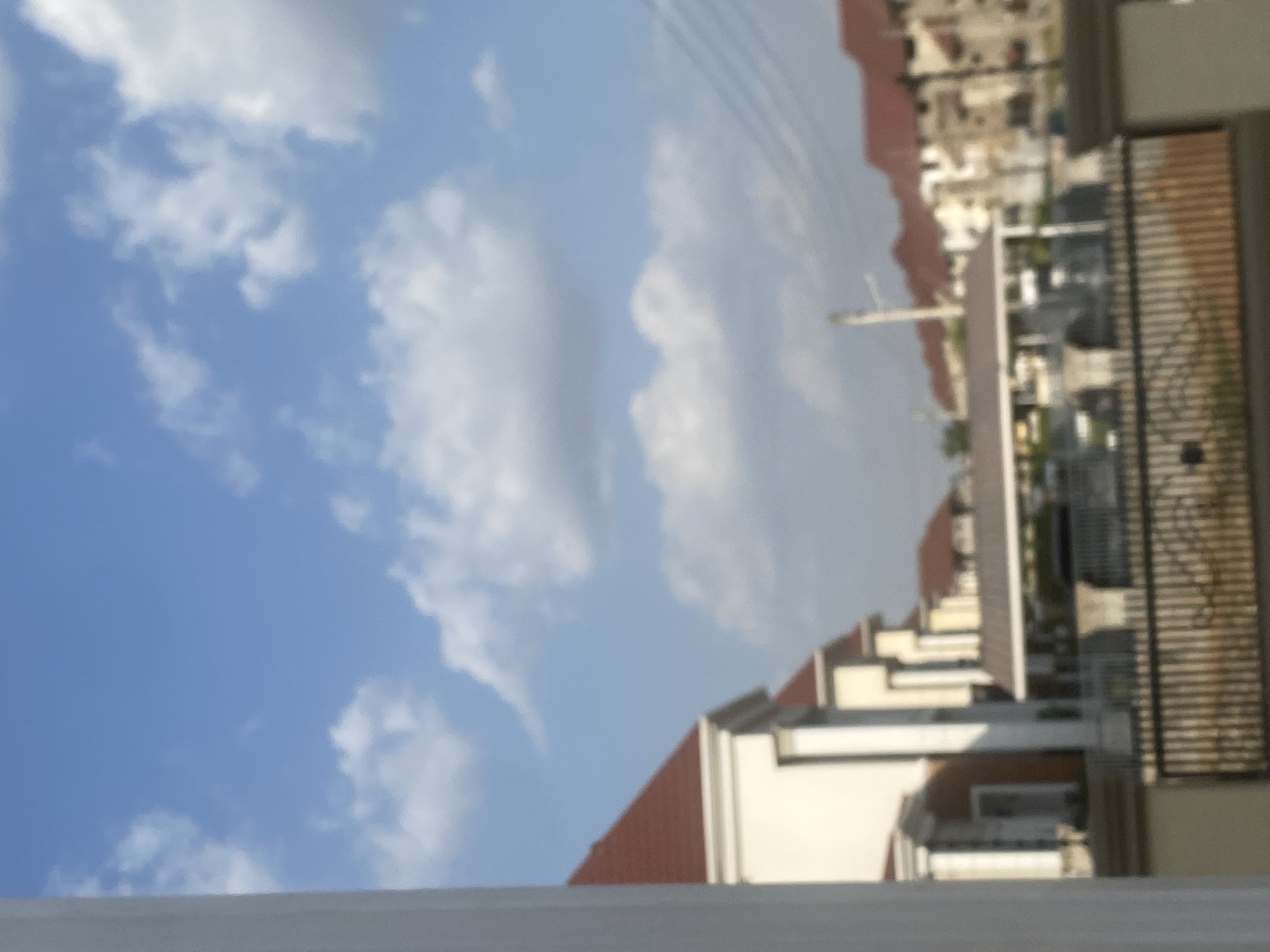 The image depicts a daytime view of buildings and sky in Iddo Tudunwada, Nigeria. The sky is blue with scattered white clouds. In the foreground, a white parapet is visible. Moving towards the right, are the buildings with red tile roofs, white walls, and dark balcony railings. In the distant background, more buildings are visible, appearing denser and further away. A utility pole extends upward into the sky. The image is slightly blurry.