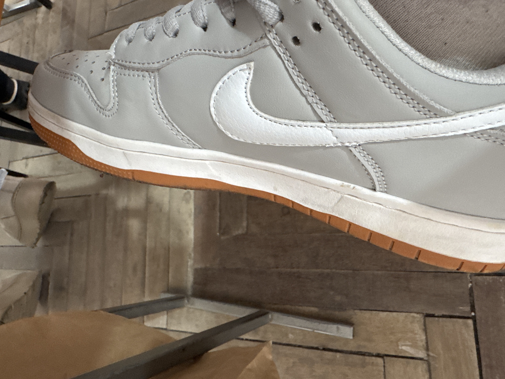 A close-up shot focuses on the side of a light grey Nike Dunk Low sneaker with a white swoosh and a gum sole. The shoe sits on a surface of wood planks. A metal table leg and a brown object, potentially cardboard, are partially visible in the foreground. The scene appears to be indoors. There is no information about the location in Yerevan, Armenia.