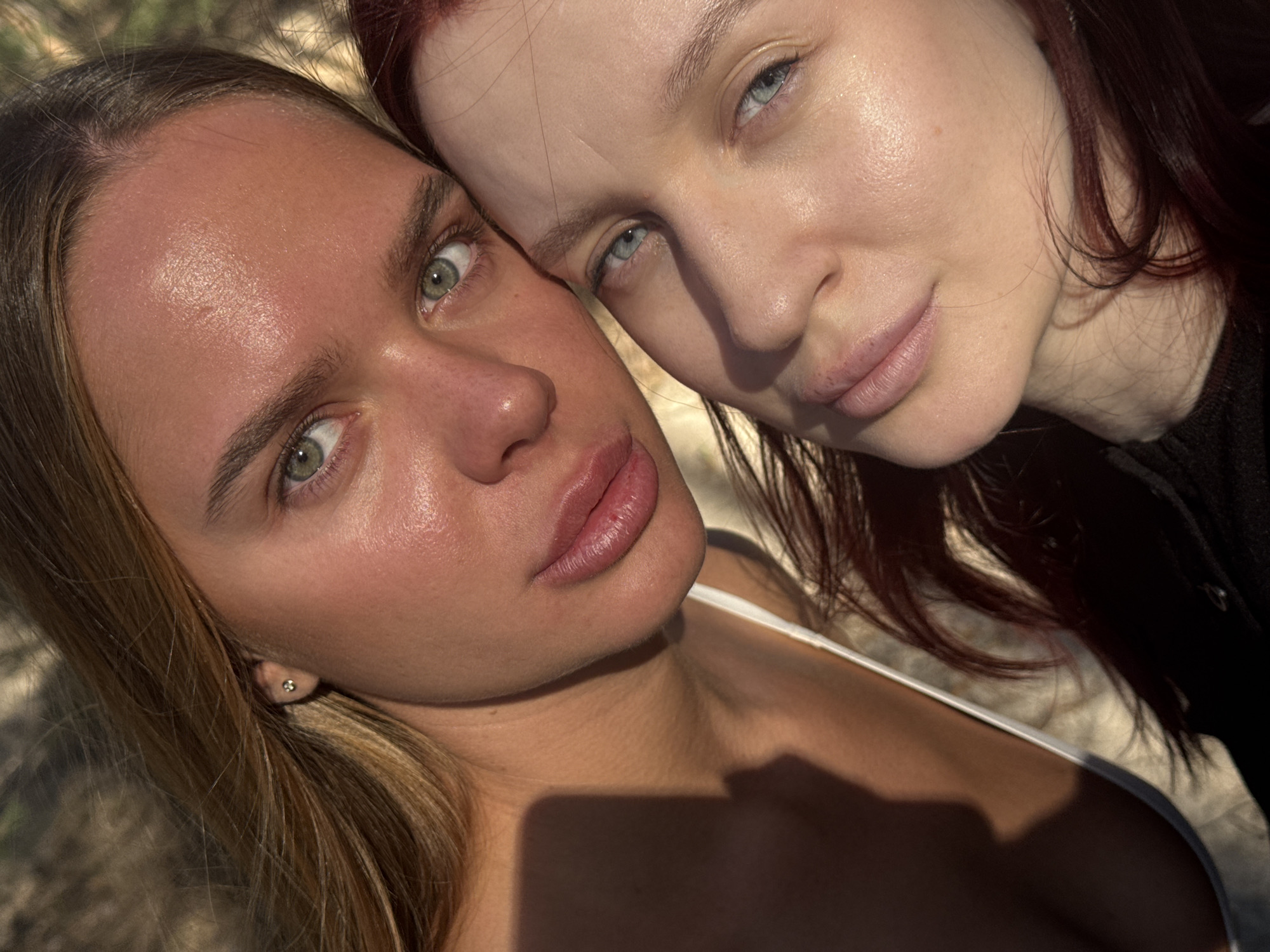 Two women are closely posed together in an outdoor setting. The woman on the left has light brown to blonde hair, green eyes, and a fair complexion. She is wearing a white top. The woman on the right has reddish-brown hair, blue eyes, and a very fair complexion. She is wearing a dark-colored top. Their faces are close together, suggesting an intimate relationship. The background is out of focus, appearing to be a natural setting possibly with foliage and dappled sunlight. There is no specific information to indicate the location is Nizhny Novgorod, Russia.