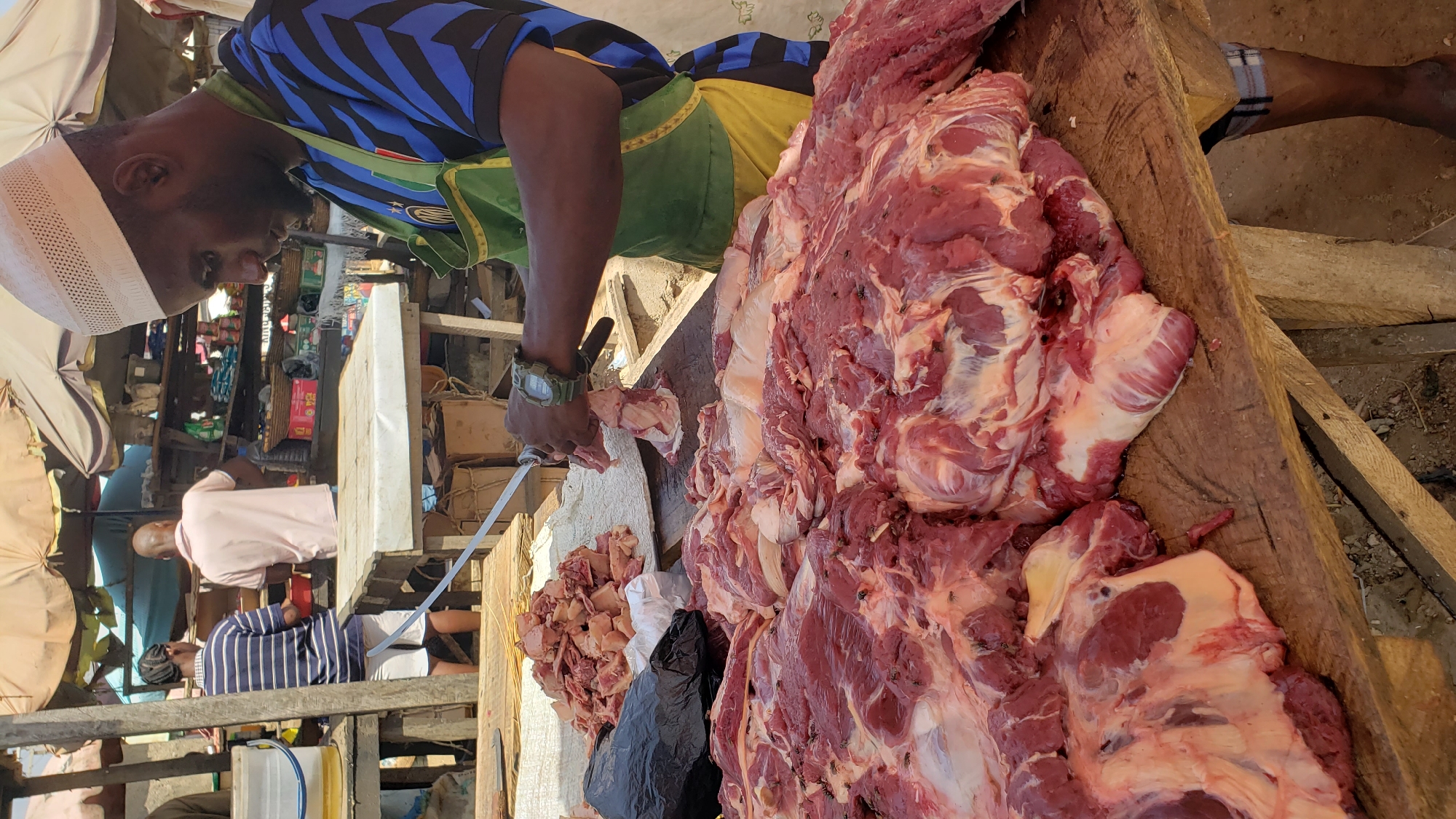 In Narayi, Nigeria, a man wearing a blue and black striped shirt, yellow and green trim, and a white kufi is cutting raw meat on a wooden surface. The meat is large and has visible veins, fat, and flies. Next to the large cut of meat are smaller pieces of cut meat and a black plastic bag. The man is using a long, thin knife with a white handle. He is wearing a wristwatch on his left wrist. Other people can be seen in the background of an outdoor market setting.