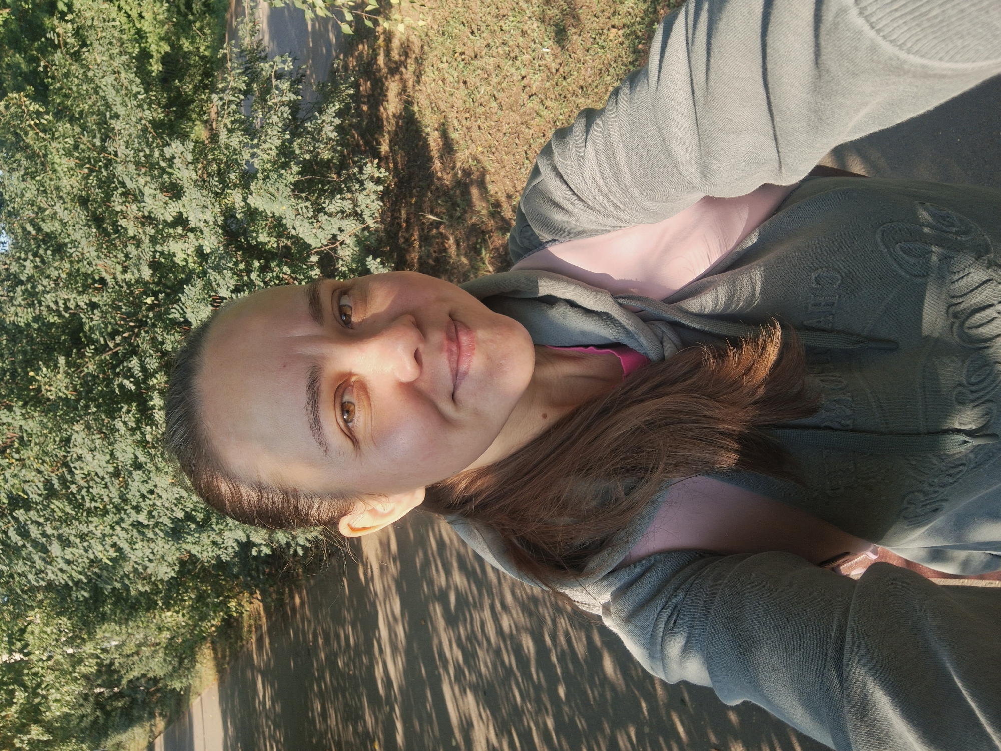 A selfie is taken by a woman with brown hair pulled back in a ponytail, wearing a grey hooded sweatshirt over a pink shirt. She has brown eyes and is smiling. The background shows a paved path with dappled sunlight and shadows, bordered by dense green foliage. The scene appears to be outdoors in a park or along a trail, possibly in or near the city of Elovo, Russia, given the sunlight and vegetation.