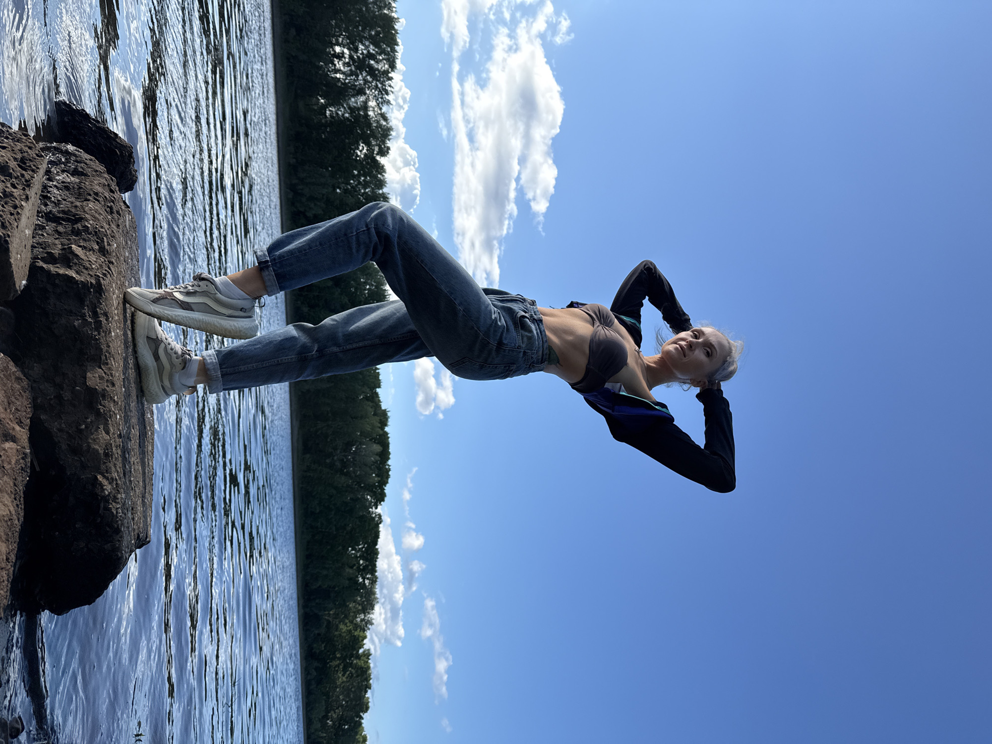 A woman stands on a large rock in the Kama River in Chaikovsky, Russia. She is wearing a dark sports bra, blue jeans, and white sneakers. Her arms are behind her head, and she is looking up at the sky. The sky is blue with some white clouds. There is a line of trees in the background. The water is calm with some ripples. The rock she is standing on is dark and rough.