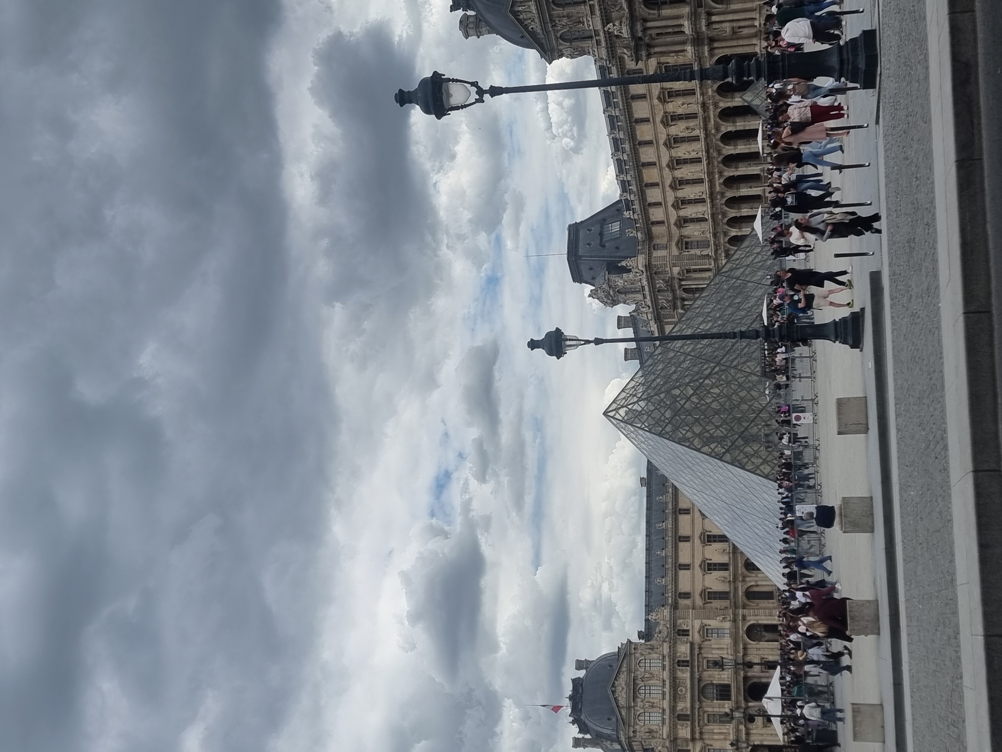 The image captures a bustling daytime scene at the Louvre Museum in Paris, France. The iconic glass Louvre Pyramid stands prominently towards the right, reflecting the cloudy sky. Surrounding the pyramid are the grand, historic buildings of the Louvre Museum, characterized by their ornate classical architecture, numerous windows, and decorative detailing.
Numerous people, presumably tourists, are visible across the square. Many are gathered in what appear to be queues, suggesting they are waiting to enter the museum or access its facilities. Others are seen walking around the area. Two tall, dark lampposts with classical designs are strategically placed, one in the mid-ground and another further back, adding to the Parisian aesthetic.
The sky is heavily overcast with prominent grey clouds, although some patches of brighter sky are visible. Despite the cloud cover, it is clearly daytime, and the scene is well-lit by natural light, casting no strong shadows. Small concrete barriers or bollards are present on the ground, likely used for crowd management. A small flag, possibly the French flag, can be seen flying from one of the museum buildings. No legible text is visible in the image. The overall atmosphere is one of a busy, popular tourist destination.