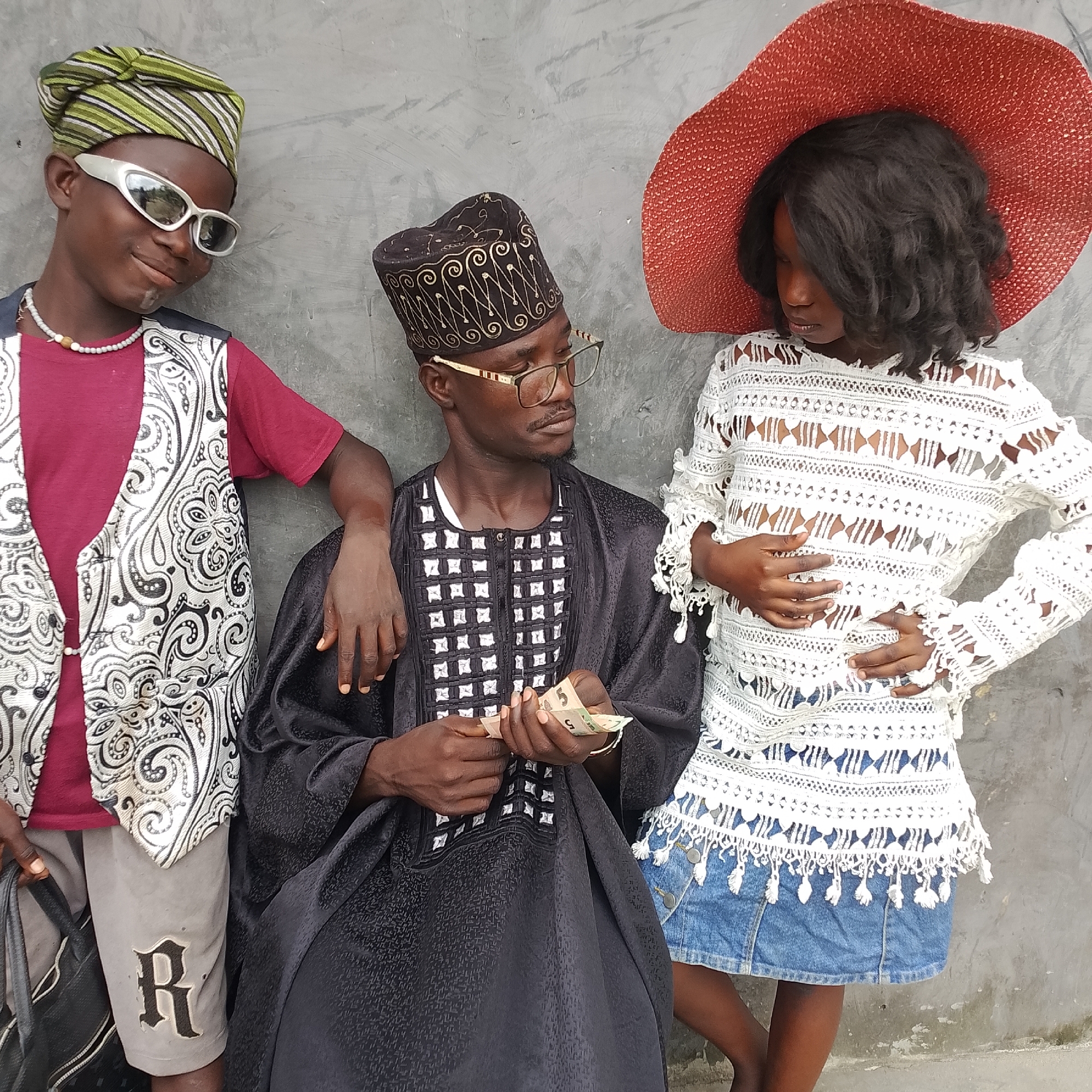 Three individuals stand against a grey, textured wall, suggestive of an urban outdoor environment in Ado, Nigeria. On the left, a young male wears a green and white striped headwrap, white sunglasses, a pearl necklace, a maroon t-shirt, and a patterned vest over grey shorts with an emblem on the left leg. He carries a black bag. In the center, a man in a black, embroidered cap and glasses holds Nigerian Naira banknotes, counting them. He is dressed in a traditional black garment. On the right, a young female, possibly a child, wears a wide-brimmed red hat and a white, intricately patterned lace top with dangling fringes, paired with a denim skirt. The overall scene appears to be a posed photograph capturing the individuals in their attire.