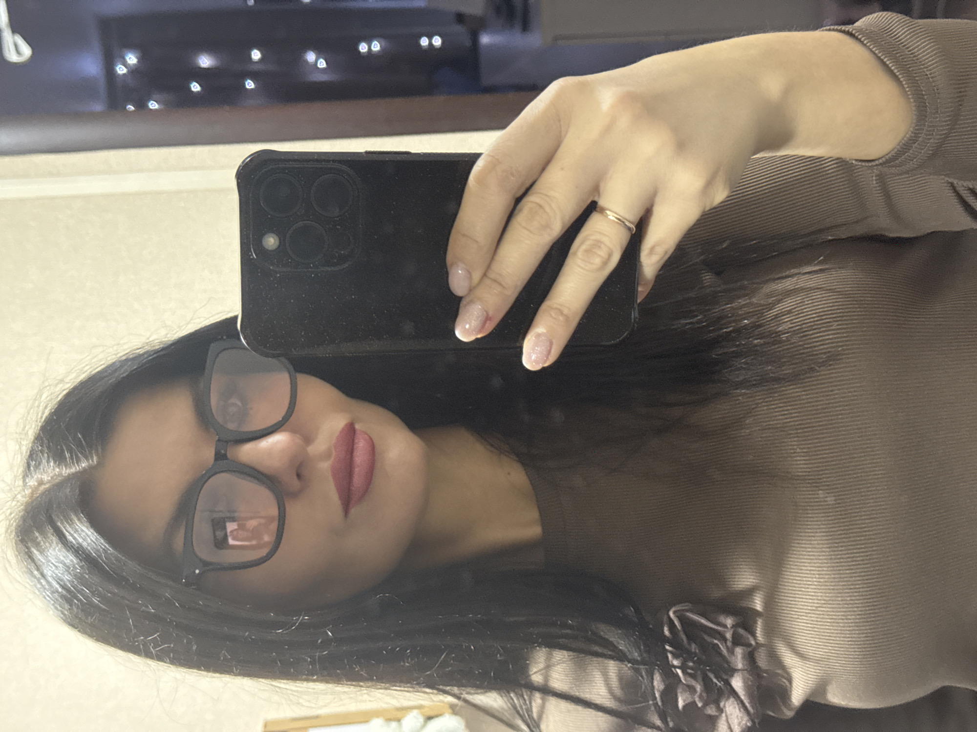 A woman with long, dark hair and wearing eyeglasses is taking a selfie in a mirror. She is holding a black smartphone with three camera lenses. She is wearing a brown, ribbed sweater. A gold ring is visible on her ring finger. The reflection in the mirror shows a window with blurred lights. The location is implied to be Chaikovsky, Russia.