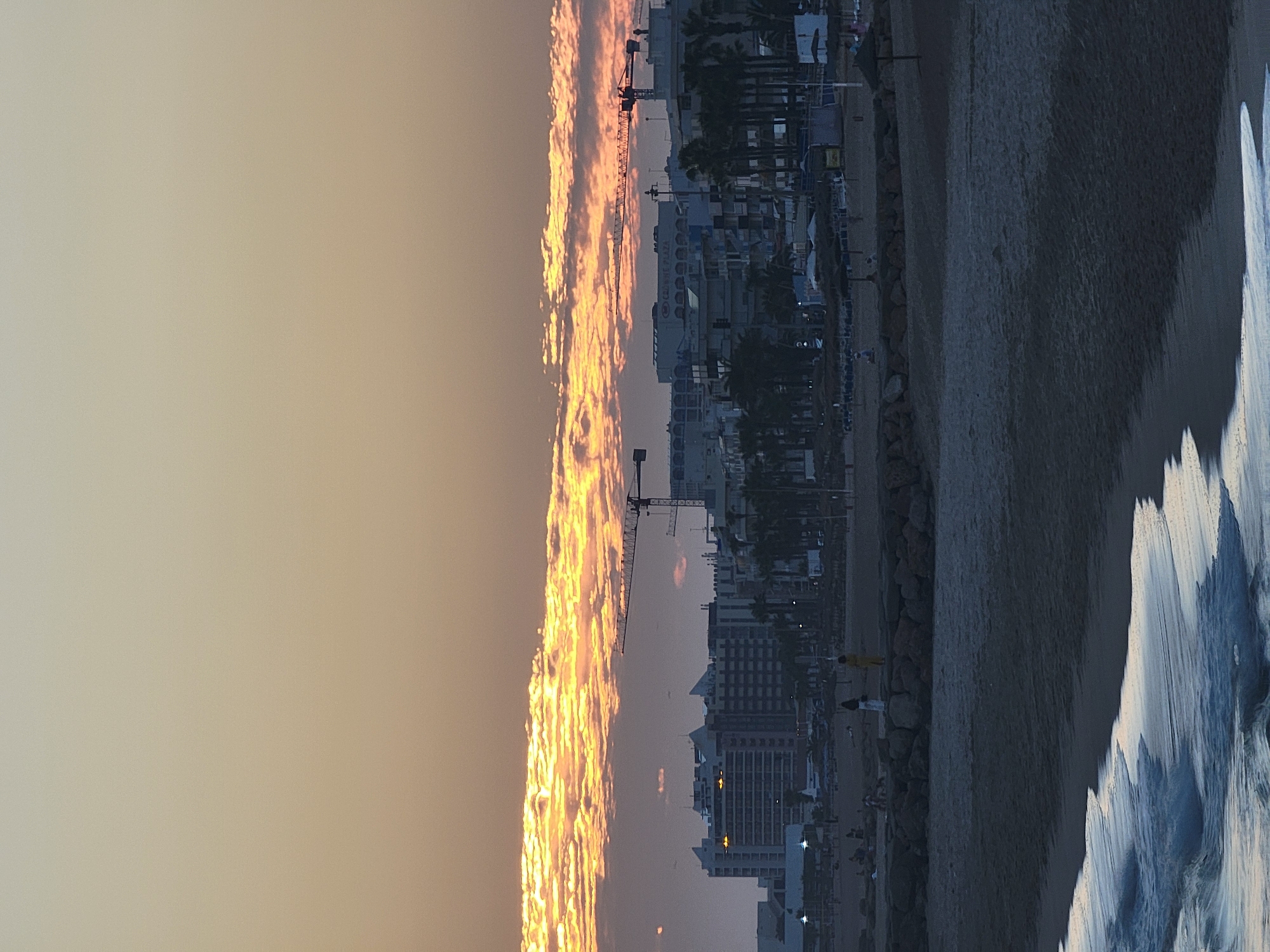This is a vertical, medium-range, eye-level shot of a beach and cityscape in Quarteira, Portugal, at sunset. The sky dominates the top half of the frame with a gradient of pale yellow and soft orange hues. A long, thin band of clouds stretches horizontally across the middle of the sky, illuminated by the setting sun with bright orange and yellow light.
Below the sky, the middle ground features a cityscape with several tall buildings, including a prominent building with the name "CROWNE PLAZA" visible. Two construction cranes are positioned in front of and above the buildings. Palm trees line the edge of the cityscape, adding a tropical element.
The foreground is a sandy beach, with the edge of the water visible at the bottom left. Gentle waves are breaking on the shore, with white foam and dark water. There are scattered rocks forming a low wall along the beach in the middle ground. A few people are visible on the beach, including two women standing on the rocky wall. The overall atmosphere is serene, with the soft light of sunset casting a warm glow over the scene.