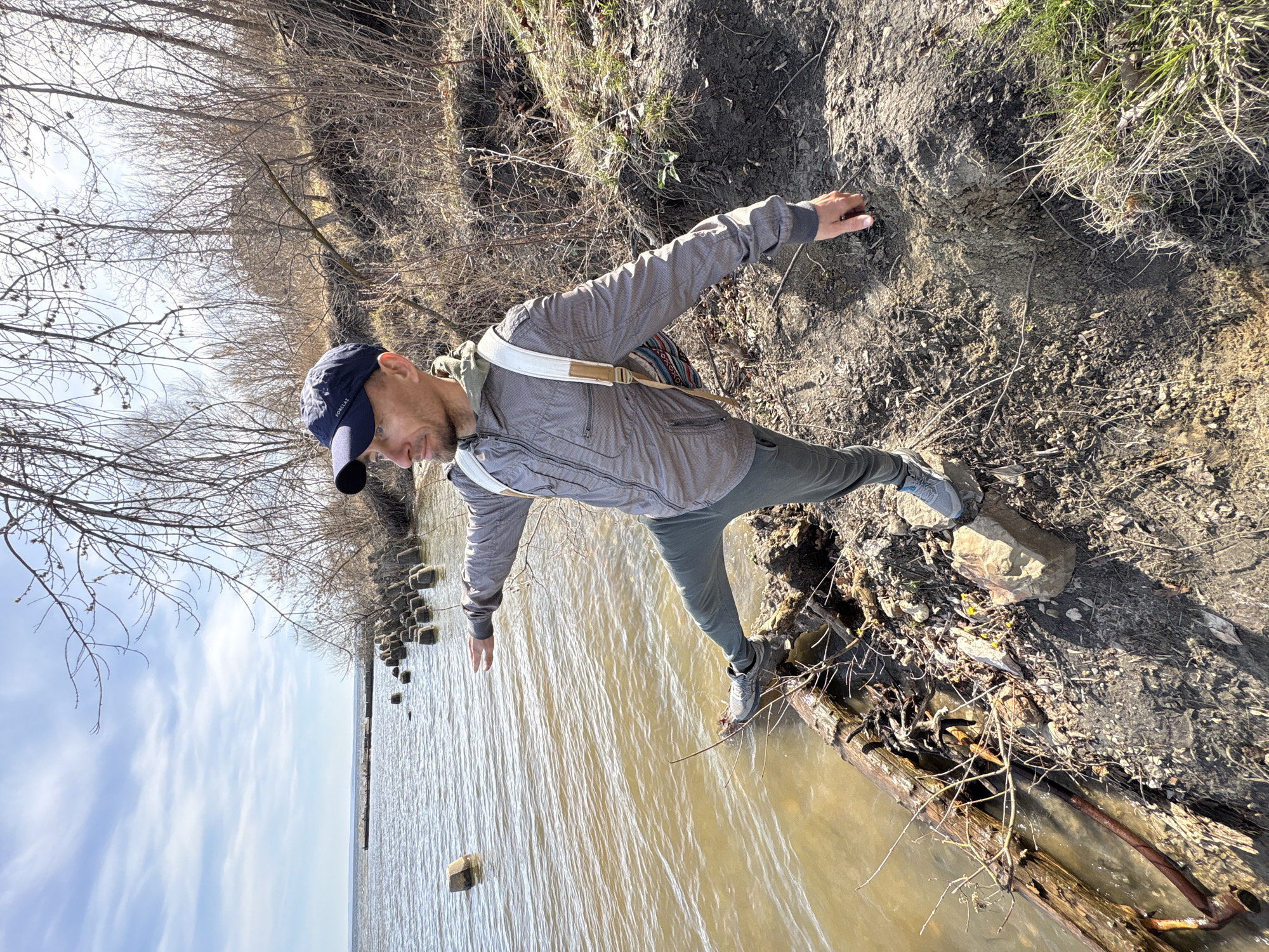 A man is depicted in an outdoor, natural setting, balancing at the edge of a muddy riverbank. He stands with his left foot on a large, partially submerged log or wooden structure in the water, and his right foot firmly on the muddy bank. His arms are outstretched to his sides, aiding his balance as he navigates the uneven terrain.
The man is dressed in a gray, patterned long-sleeved jacket, dark trousers, and gray and blue athletic shoes. He wears a dark blue baseball cap with the brand "FORCLAZ" visible on the front, and a light-colored shoulder strap from a bag crosses his chest. His short beard is visible, and his gaze is directed downwards, focused on his footing.
The scene features a wide body of brown, rippling water, extending towards a distant horizon under a sky with scattered white, wispy clouds. Along the water's edge, several dark, square-shaped objects, possibly old wooden posts or rocks, are partially submerged. The riverbank is steep and covered with bare tree branches and dry, sparse vegetation, suggesting a late autumn, winter, or early spring season. The ground is composed of dark, loose soil and mud. Bright sunlight illuminates the scene, casting shadows and indicating daytime, likely late morning or early afternoon. The overall impression is a cautious exploration of a natural, rugged environment in Ulyanovsk, Russia.