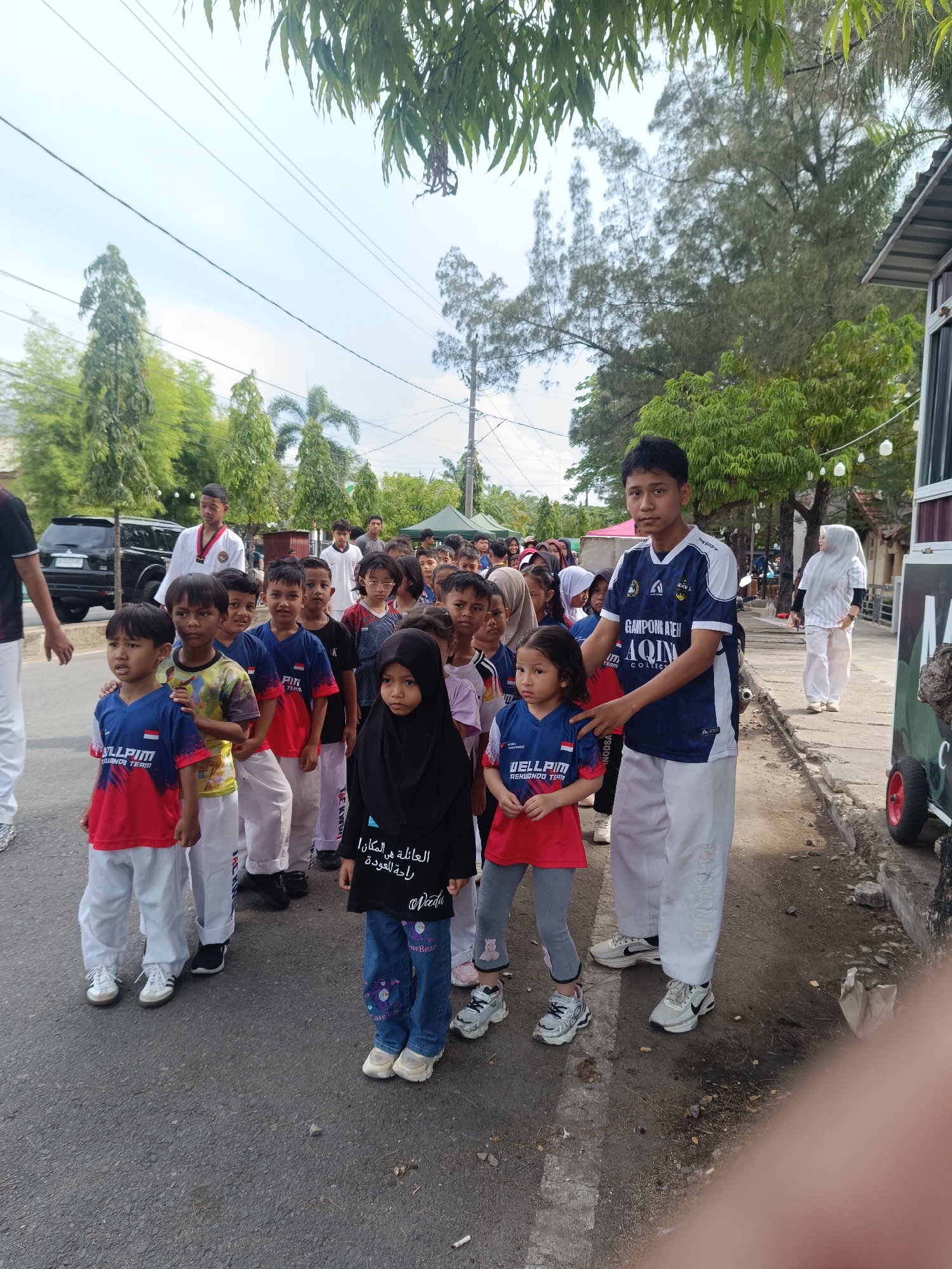 A group of children and adults are standing on a paved street in what appears to be an outdoor gathering or parade. The location is likely in Lhokseumawe, Indonesia, given the text on clothing and other visual cues. The group includes children mostly wearing matching red and blue sports shirts and white pants. An older teen or young adult is directing or assisting the children, wearing a blue and white sports shirt. A girl is wearing a black hijab. A few other adults are present, some in white taekwondo uniforms. A black SUV is parked on the side of the road. Trees and buildings line the background. The weather appears to be overcast.
