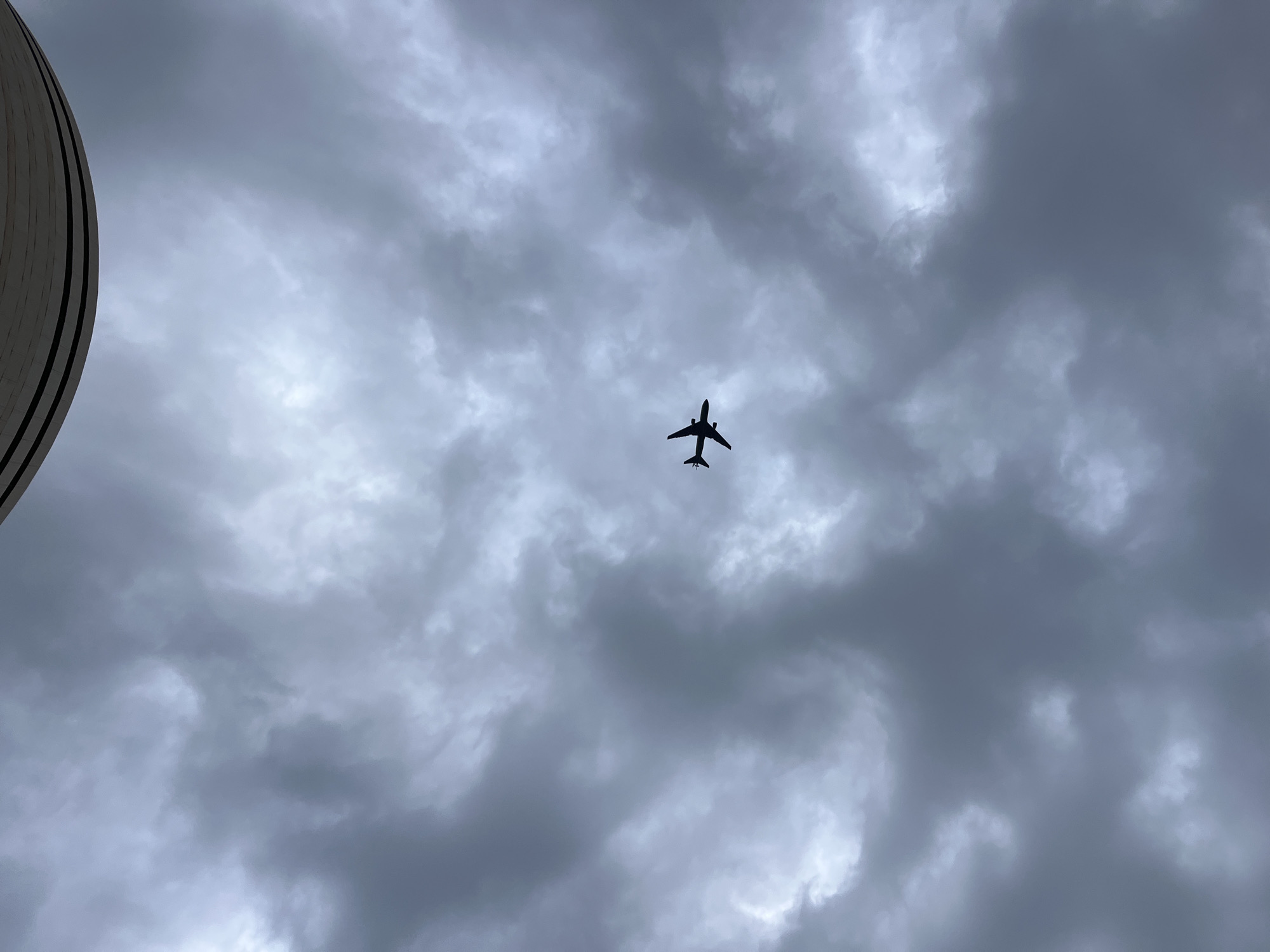 A silhouetted airplane is flying in the sky, which is covered by gray storm clouds. In the bottom left corner of the image is a rounded building structure, with the light-colored building covered with dark horizontal stripes. I have no information on the exact location in the city of Tel-Aviv, Israel.