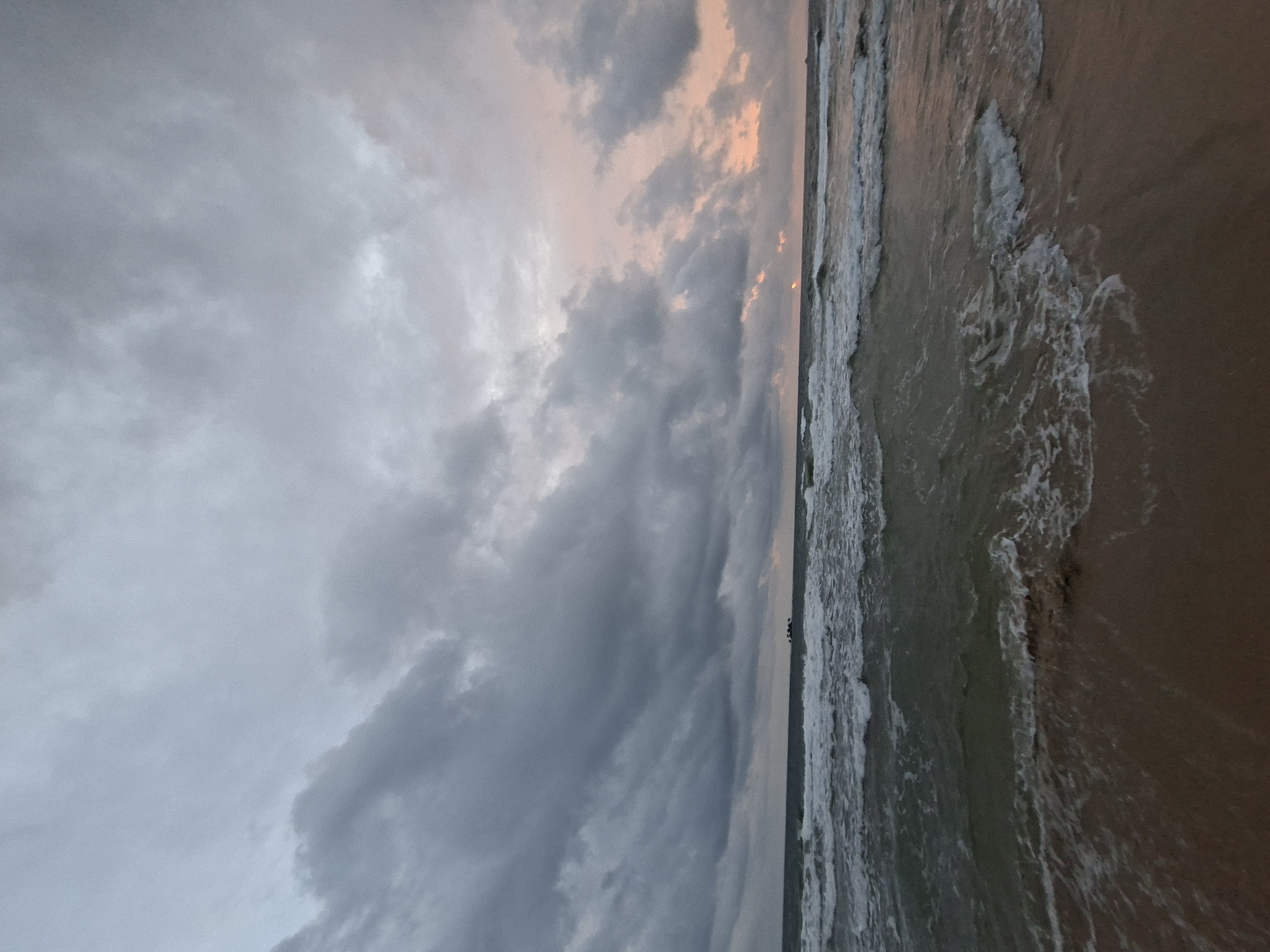 A wide-angle, low-angle shot captures an overcast sky over the ocean and a beach on Phú Quốc, Vietnam. The sky is filled with dense, dark gray clouds, with a faint blush of pink and orange peeking through near the horizon on the right, suggesting the approach of dawn or dusk. The ocean is moderately rough, with white-capped waves rolling towards the shore. A dark, low-lying mass, possibly a small island or a cluster of rocks, is visible on the horizon in the distance, slightly to the left of center. The beach in the foreground is composed of wet sand, with the incoming waves washing over it, creating foamy patterns. The overall atmosphere is moody and dramatic, with the muted colors and cloudy sky creating a sense of anticipation or stillness. There are no people or specific structures visible in the image.