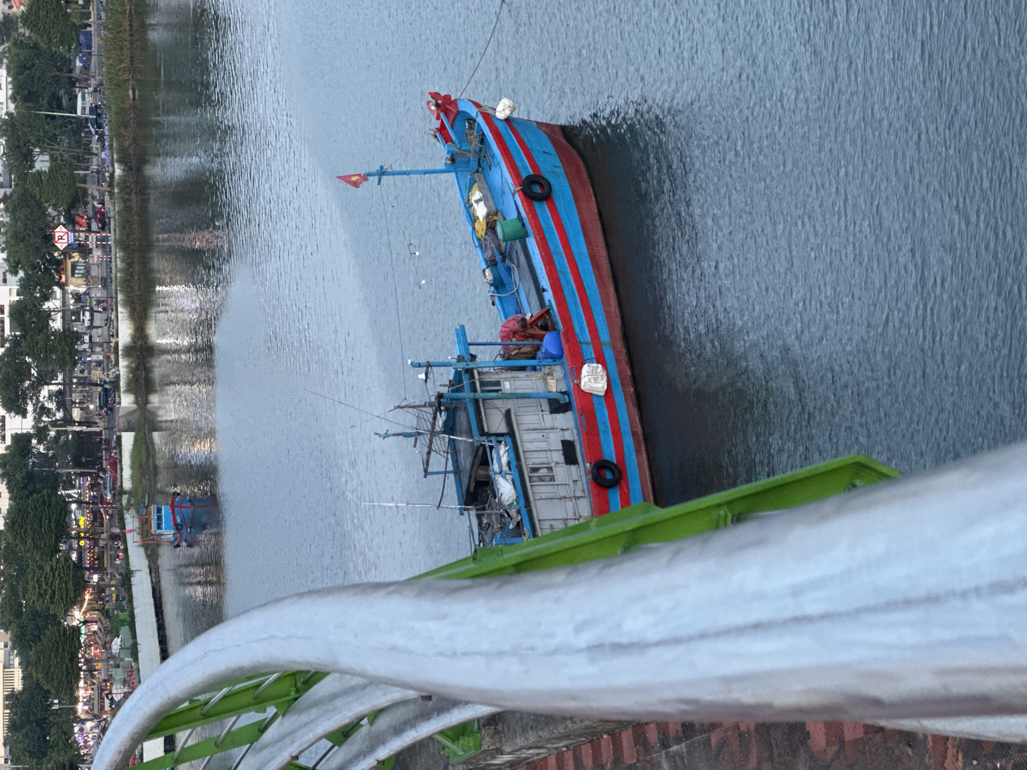 A view from a bridge in Đà Nẵng, Vietnam, looking down at a river with a fishing boat docked near the bank. The boat is painted with blue and red stripes and has a Vietnamese flag. A person can be seen onboard. The city skyline with buildings, trees, and streetlights lines the opposite riverbank. The bridge railing is visible in the foreground.