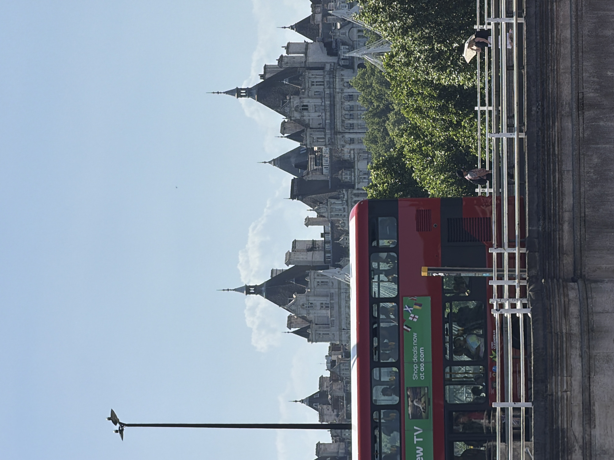 This photograph depicts a red double-decker bus traveling on a bridge in London, United Kingdom. The bus, prominently displayed, carries passengers. The building behind the bus appears to be historical. The scene is set on a clear day, with a blue sky. A lamp post is visible in the foreground. People can be seen on the bridge.