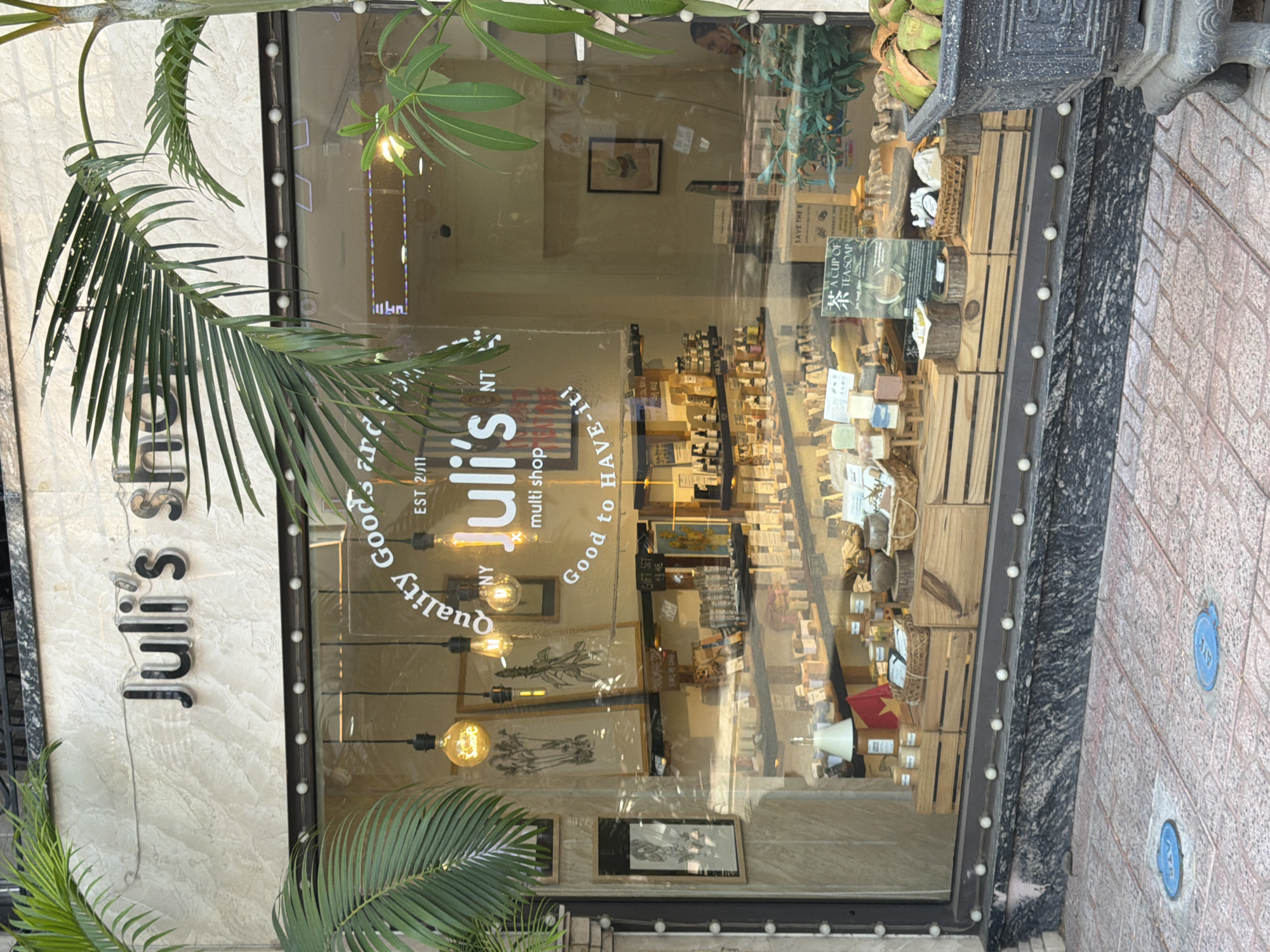 A street-level, eye-level, outdoor, medium shot shows the storefront of "Juli's shop" in Nha Trang Ward, Vietnam. The shop's facade features a marble-like material with the shop's name in large, black, stylized letters above a glass window. The window displays various items, including soaps, wooden crates, and decorative plants. Inside, shelves are stocked with bottles and packaged goods, suggesting a retail environment. A person is partially visible in the background through the window. Palm trees frame the storefront, and the foreground shows a tiled sidewalk with circular blue utility covers. The lighting suggests it is daytime, possibly late afternoon given the warm glow of the Edison-style bulbs hanging in the window.