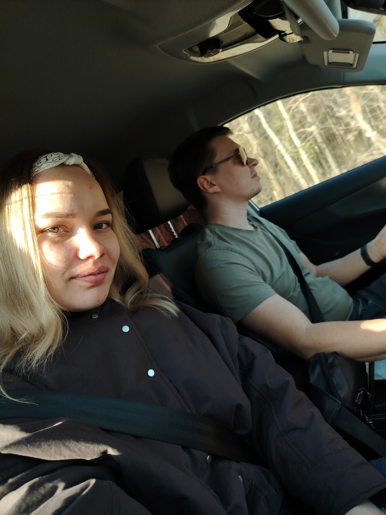 A woman and a man are sitting in the front seats of a car. The woman is in the passenger seat and is wearing a black jacket, a black seat belt, and a white bandana. The man is in the driver's seat and is wearing a green t-shirt and sunglasses. He is holding the steering wheel. Both are wearing seatbelts. The background shows trees visible through the car window, suggesting they are driving outside of the city. The location is Chaikovsky, Russia.