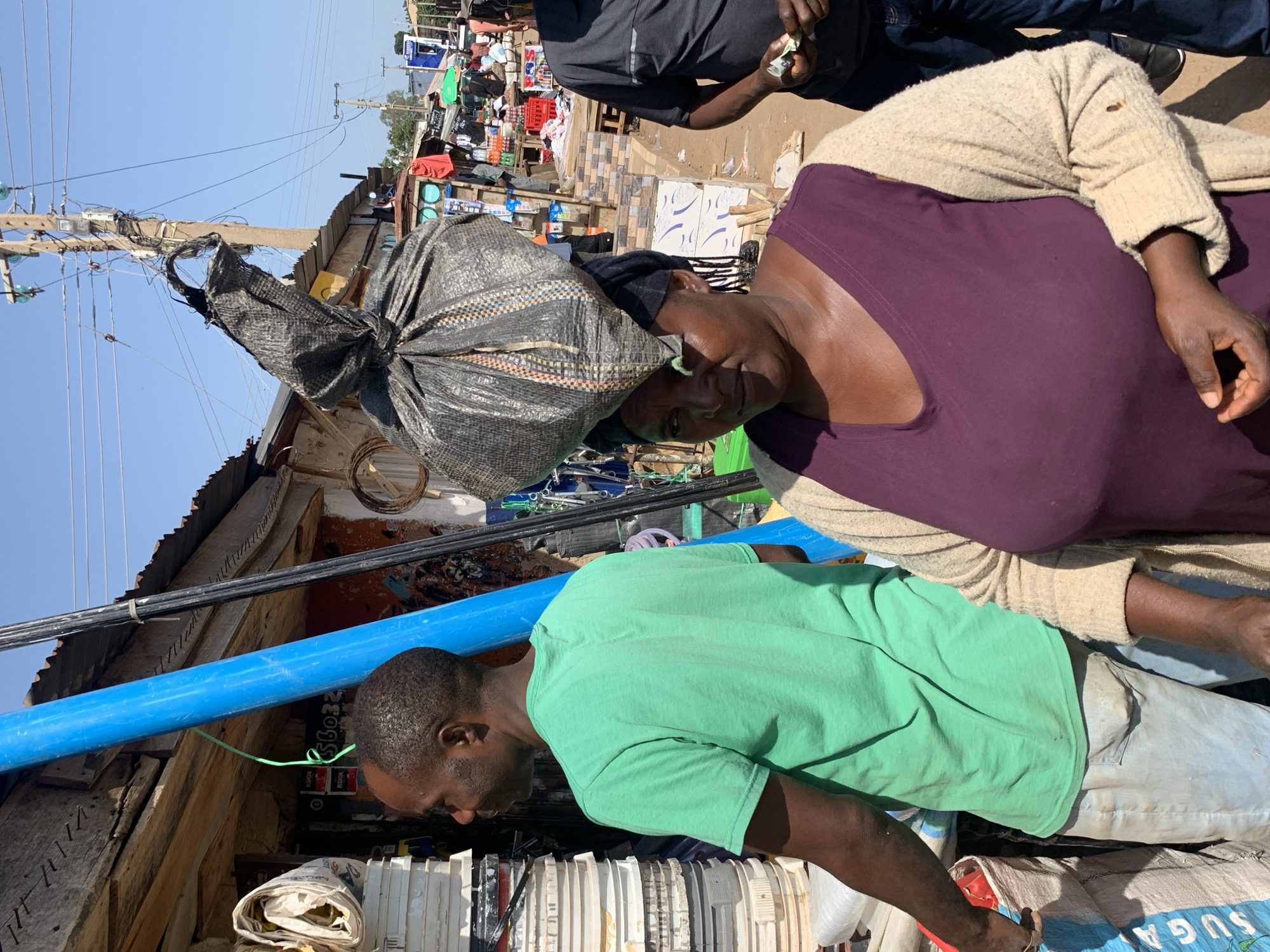 The image depicts a bustling outdoor market scene in Gura Top, Nigeria. In the foreground, a woman with a large, dark-colored bag balanced on her head is looking down. She is wearing a purple top and a light-colored cardigan. Beside her, a man in a green t-shirt is also looking downwards, his hands out of view. They appear to be in a crowded area with various stalls and goods.
Behind the main subjects, the market is filled with an assortment of items, including what appear to be crates of drinks, packaged goods, and possibly textiles. Power lines and utility poles crisscross the sky, suggesting an urban or semi-urban environment. The sunlight indicates it is daytime, and the clear sky suggests fair weather. The overall impression is one of a busy commercial hub.
The scene is rich with detail suggesting economic activity. Bags of goods are piled up, and there are signs of everyday commerce. The people are dressed in casual clothing, fitting for a market environment. There is a sense of everyday life and work in progress. Some text is visible on blue bags in the lower right corner, but it is partially obscured and difficult to read clearly. The orientation of the image is rotated, with the subjects appearing to be standing at an angle.