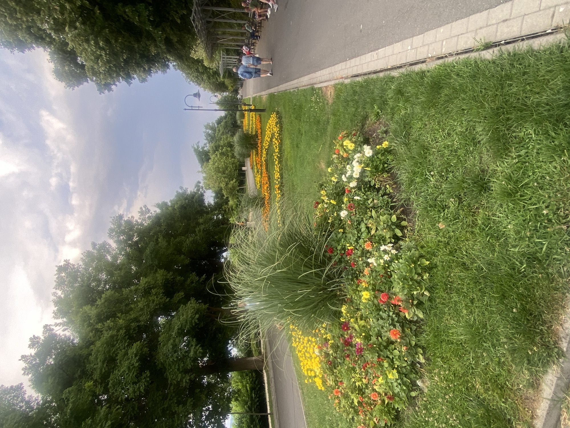 The image depicts a park scene with lush green trees and a paved walkway. Several people are visible walking and sitting along the path. The walkway is bordered by a well-maintained lawn and flower beds bursting with colorful flowers in shades of yellow, orange, red, and white. A tall, ornamental grass adds texture to the landscaping. A lamppost stands near the walkway, suggesting it's an urban park or garden. The sky is partly cloudy, indicating it's daytime, likely late morning or afternoon. The overall atmosphere appears relaxed and pleasant, suggesting a leisurely outing. The location is identified as Burgas, Bulgaria, placing the scene in a coastal city in Southeastern Europe. There is no visible text in the image.