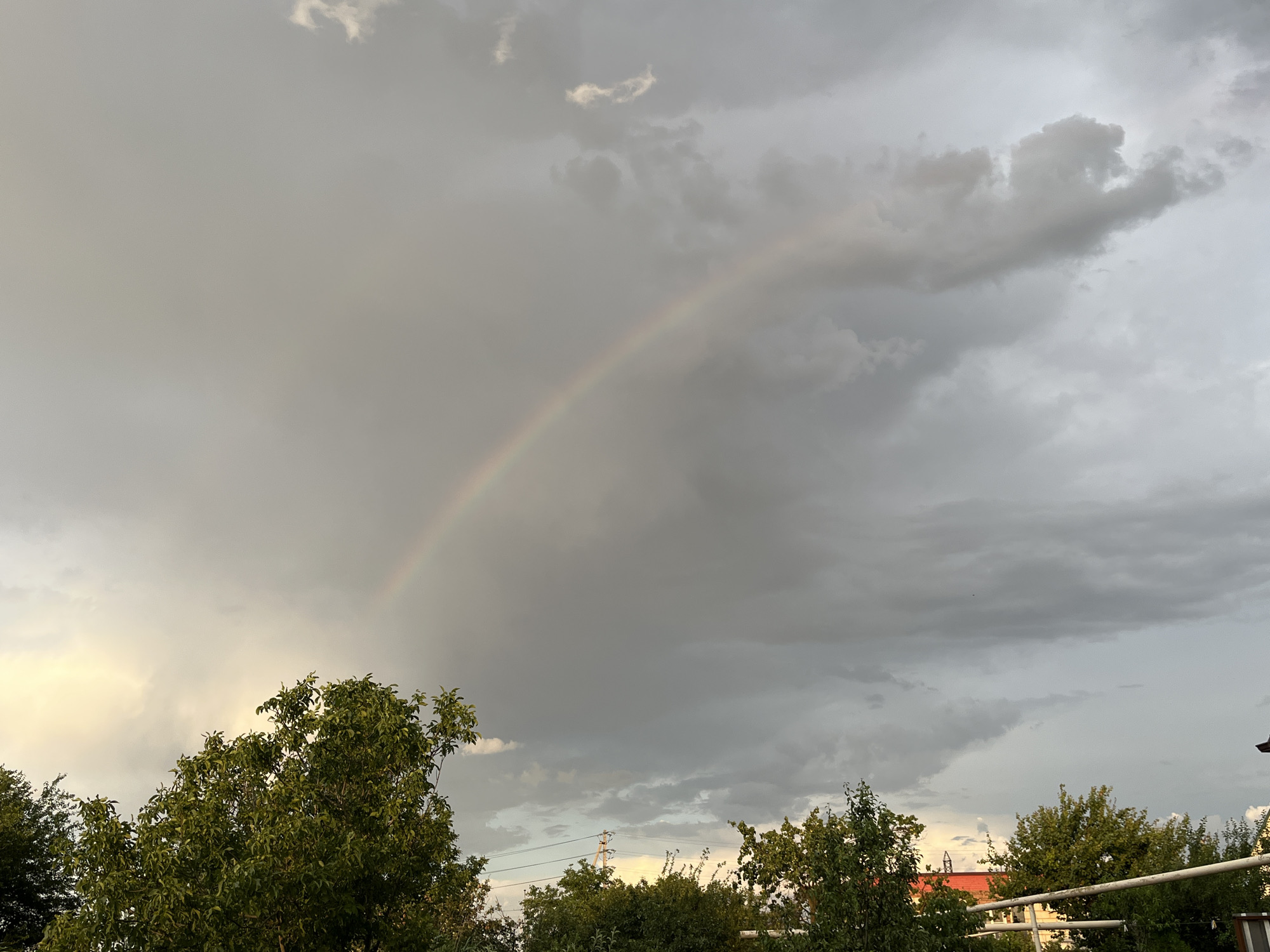 The image depicts a cloudy sky with a rainbow arching across it. The rainbow is faint, suggesting it may be early morning or late evening. Dark, heavy clouds dominate the sky, with some patches of lighter, thinner clouds. The lower part of the image shows a line of trees with lush green foliage. In the background, behind the trees, are hints of buildings with red roofs and power lines, suggesting an urban or suburban environment. The overall impression is one of a dramatic sky after or during a rain shower. The location of Krasnyye Barrikady, Russia, indicates a specific geographic context for this natural phenomenon.
