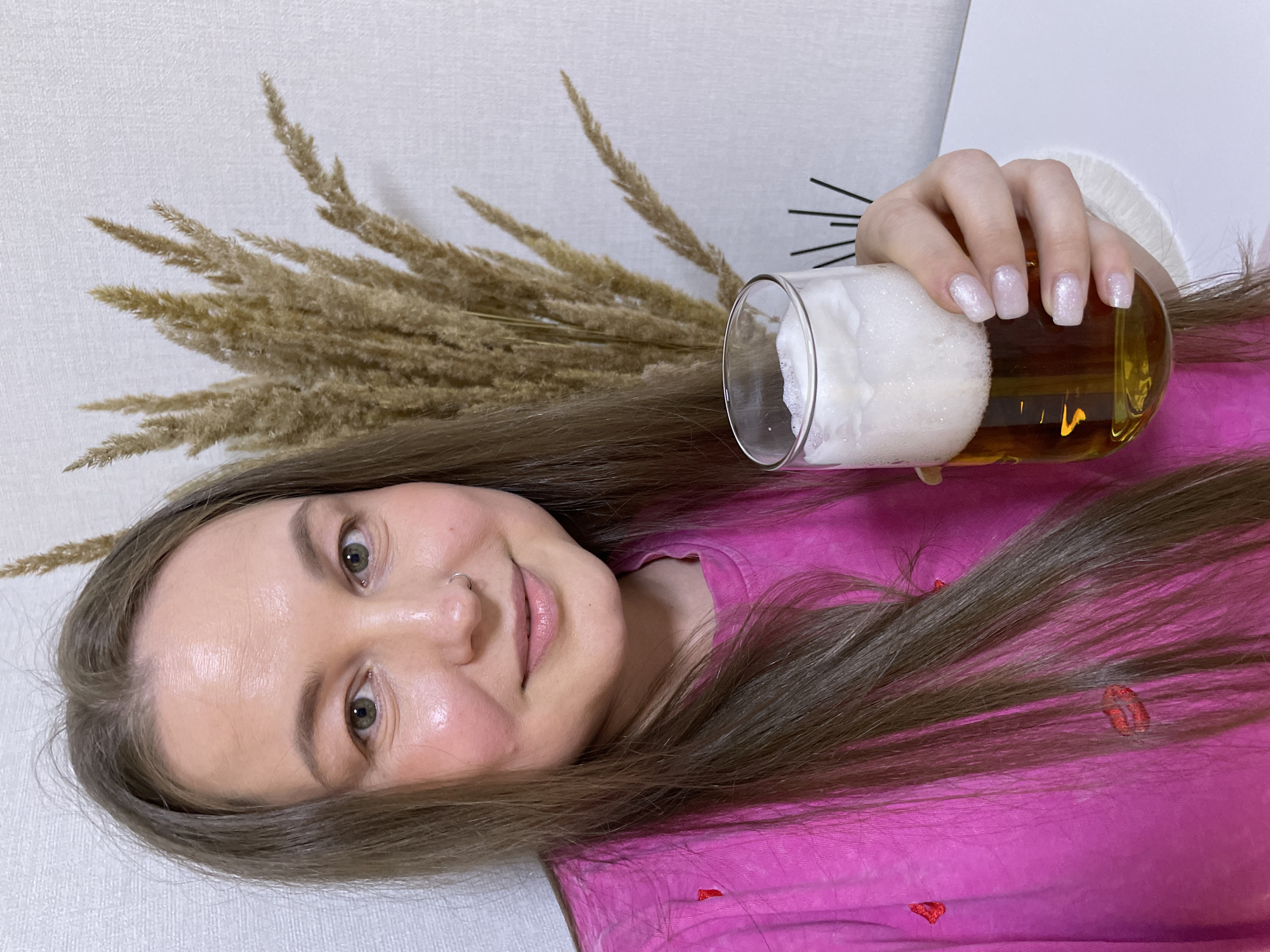 The image depicts an adult female indoors in the city of Pervomaysky, Russia. She has long, straight, medium-brown hair parted in the middle, green eyes, and a small silver ring piercing her left nostril. She is looking directly at the camera with a slight smile and light makeup, including blush and lip gloss. She is wearing a bright pink t-shirt adorned with subtle red embroidered lip motifs. Her right hand, featuring square-shaped fingernails painted with a shimmery, light, possibly iridescent polish, holds a clear glass filled with a golden-amber liquid topped by a thick layer of white foam, consistent with beer. The background consists of a light-colored, textured wall. To the right of the woman's head (viewer's left), a large arrangement of dried, feathery, light brown ornamental grasses is visible. In the lower right background, behind the glass, several thin, dark, vertical decorative sticks are partially visible.