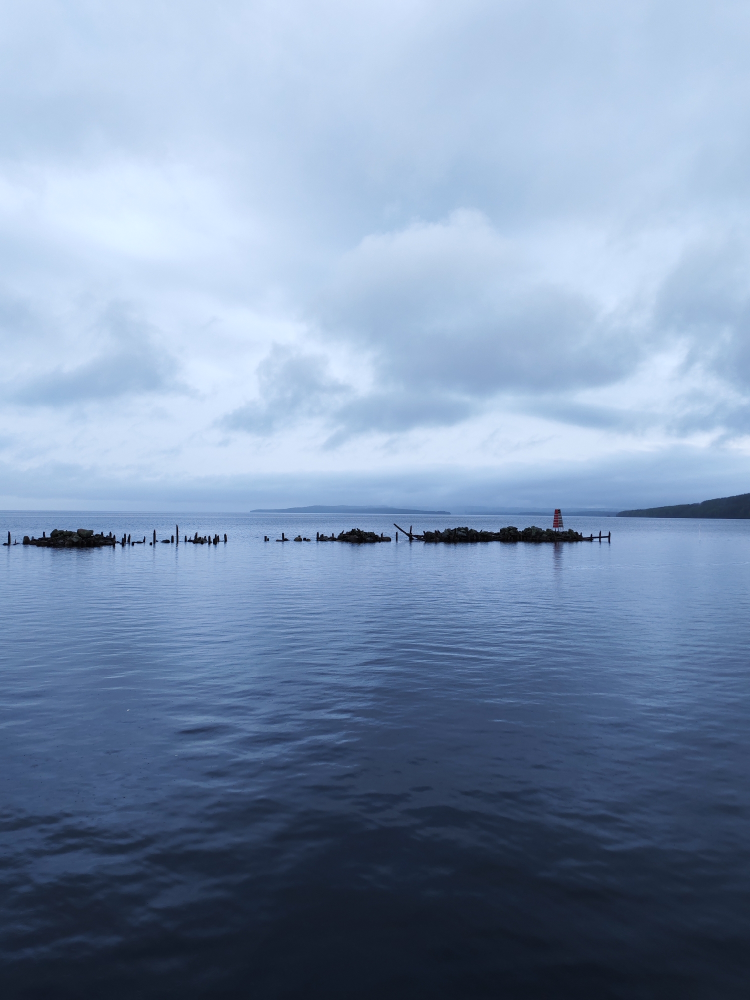 This wide-angle landscape photograph captures a serene, overcast scene in Medvezhyegorsk, Russia, likely on Lake Onega. The primary subjects are the expansive body of dark blue water, a long, low-lying man-made structure, and a heavily clouded sky.
The calm water, exhibiting subtle ripples, occupies the majority of the lower frame, its deep hues reflecting the subdued light. Stretching across the mid-ground is an artificial barrier, possibly a remnants of a pier or breakwater, composed of dark rocks and numerous weathered wooden posts protruding vertically from the water. Towards the right end of this structure, a small, distinct navigational beacon stands out with its bright red and white horizontal stripes, providing a focal point and a splash of color against the muted backdrop.
In the distance, a faint, low-lying landmass or shoreline is visible under the expansive sky. The sky itself is filled with thick, light grey and white clouds, indicating an overcast day. The diffused light contributes to a cool, tranquil, and somewhat melancholic atmosphere. No activity or people are visible, emphasizing the quiet solitude of the landscape. There is no discernible text in the image.