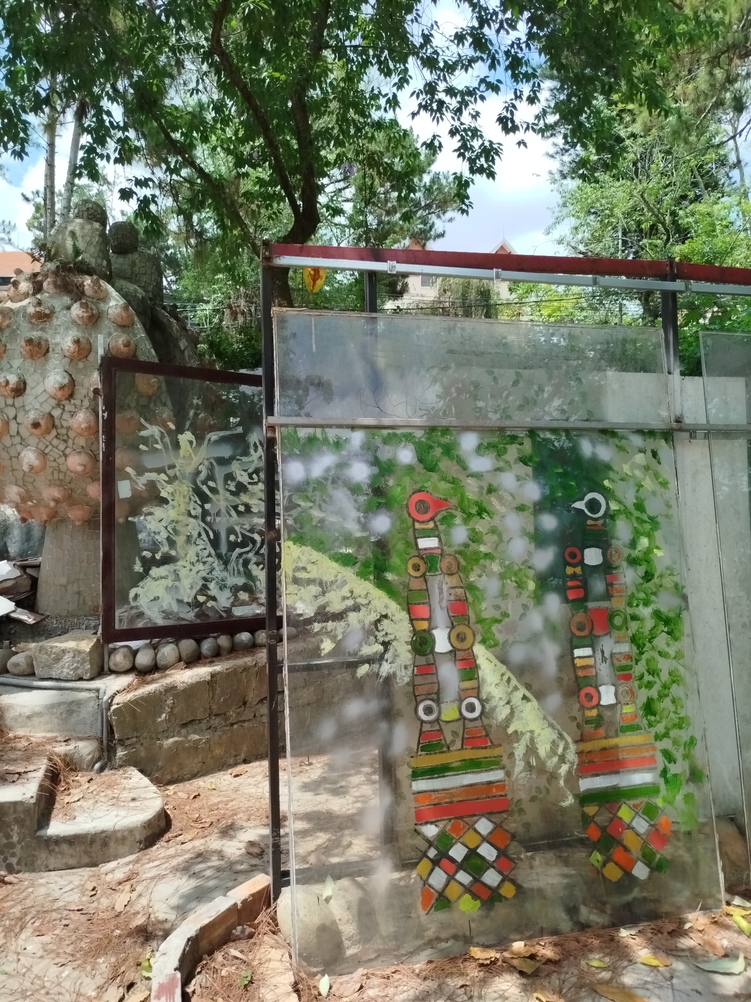 The image shows an outdoor art installation in Da Lat, Vietnam. A sculpture made of stones and embedded ceramic objects is visible in the background, to the left. In the foreground, a metal rack supports several panes of glass, some painted with abstract and stylized figures. The glass reflects the surrounding foliage. The ground is covered with fallen pine needles and the area appears to be a part of a garden or park setting, with trees in the background. The composition presents a mix of natural and man-made elements.