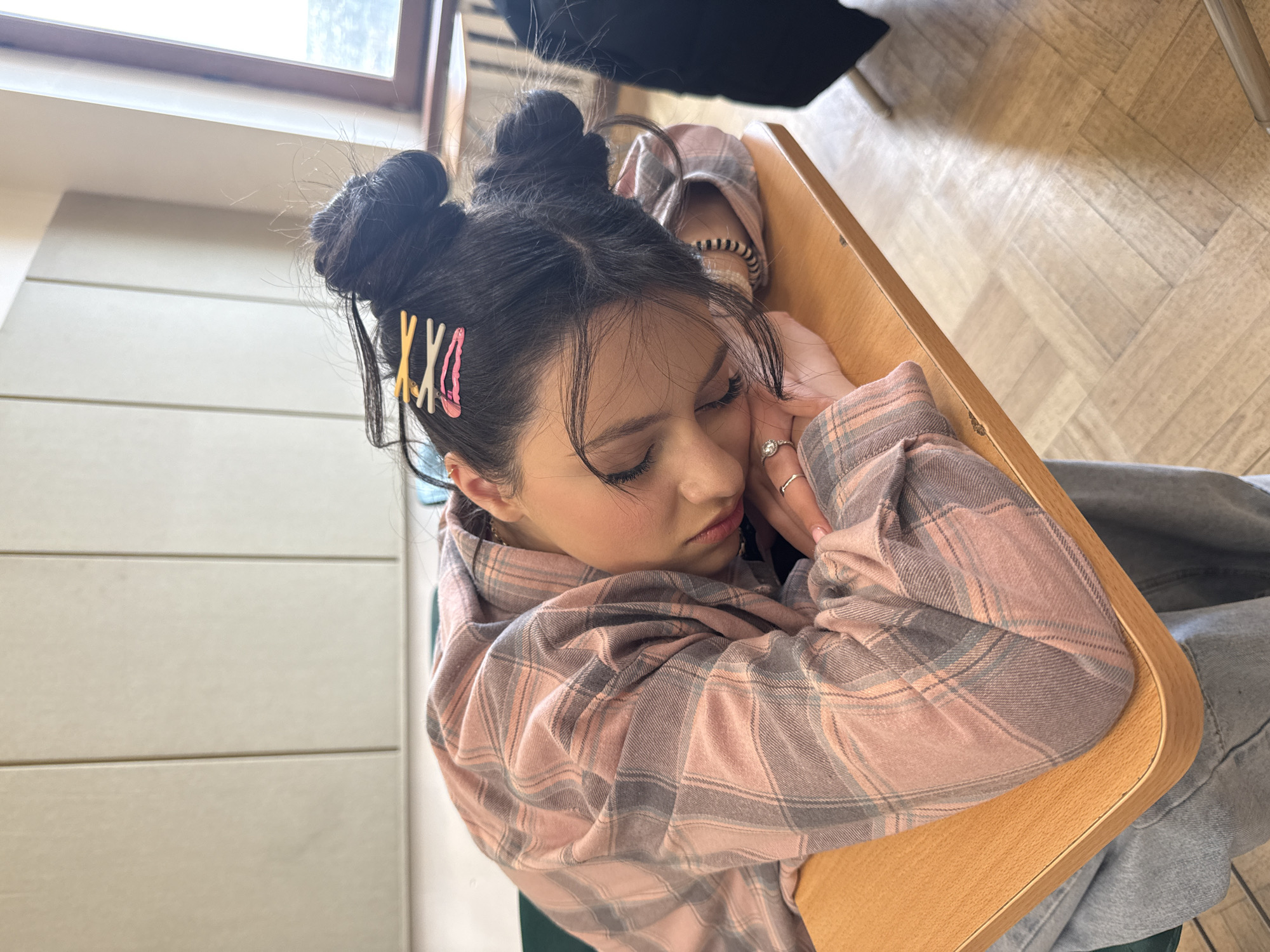 A young woman is asleep, resting her head on her crossed arms on a light-brown, wooden desk or table. She is wearing a pink and gray plaid flannel shirt and gray pants. Her dark hair is styled in two buns on top of her head, adorned with pink and yellow hair clips on the left side. She has rings on her fingers. The location appears to be indoors, possibly a classroom. The background is blurry, but there is a window and what seems to be furniture in the background. The city of Yerevan, Armenia, cannot be confirmed.