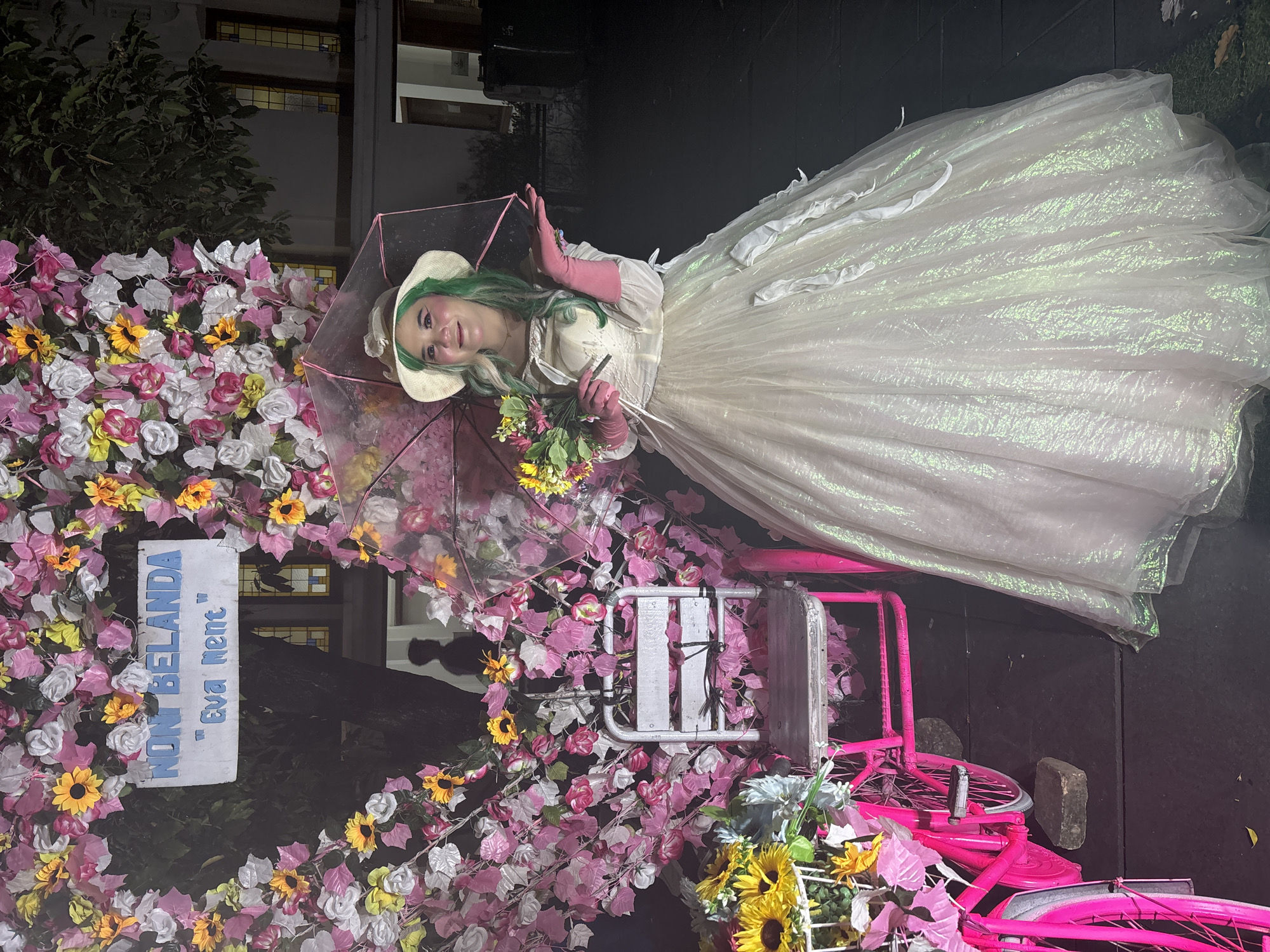 The image shows a woman dressed in a white, iridescent gown, wearing a straw hat and pink gloves, standing in front of a floral arch. She is holding a transparent umbrella and a bouquet of flowers. Behind her, a pink bicycle with a basket of flowers is visible. A sign with blue text reads "NONI BELANDA 'Eva Nent'". The scene appears to be set up outdoors at night, possibly for an event or photoshoot, with building lights and trees in the background. The location is identified as Taman Fatahillah in Kota Tua, West Jakarta, Indonesia, a historic area known for its Dutch colonial architecture.