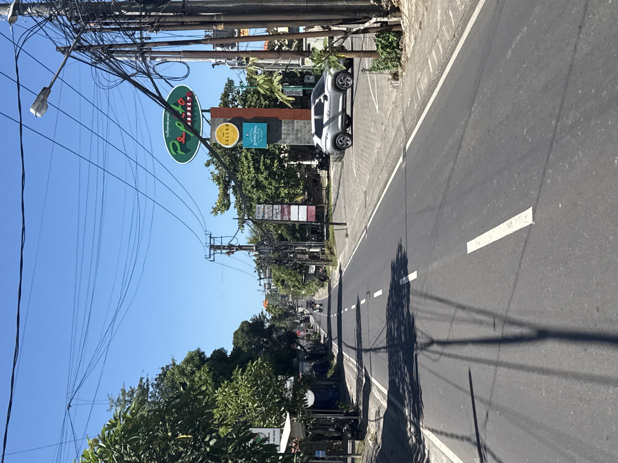 The image shows a street in Unknown, Unknown. On the left side of the street, there is a sign for "Pepito Market" and "Cellar Fine Foods & Gourmet Grocer". A white car is parked on the side of the road. Power lines and poles are visible above the street. The road has a white line marking the edge and dashed lines down the middle. Shadows of the power lines and trees are cast on the road. Trees and other vegetation are visible on both sides of the street. In the distance, other vehicles and buildings can be seen. The sky is clear and blue.