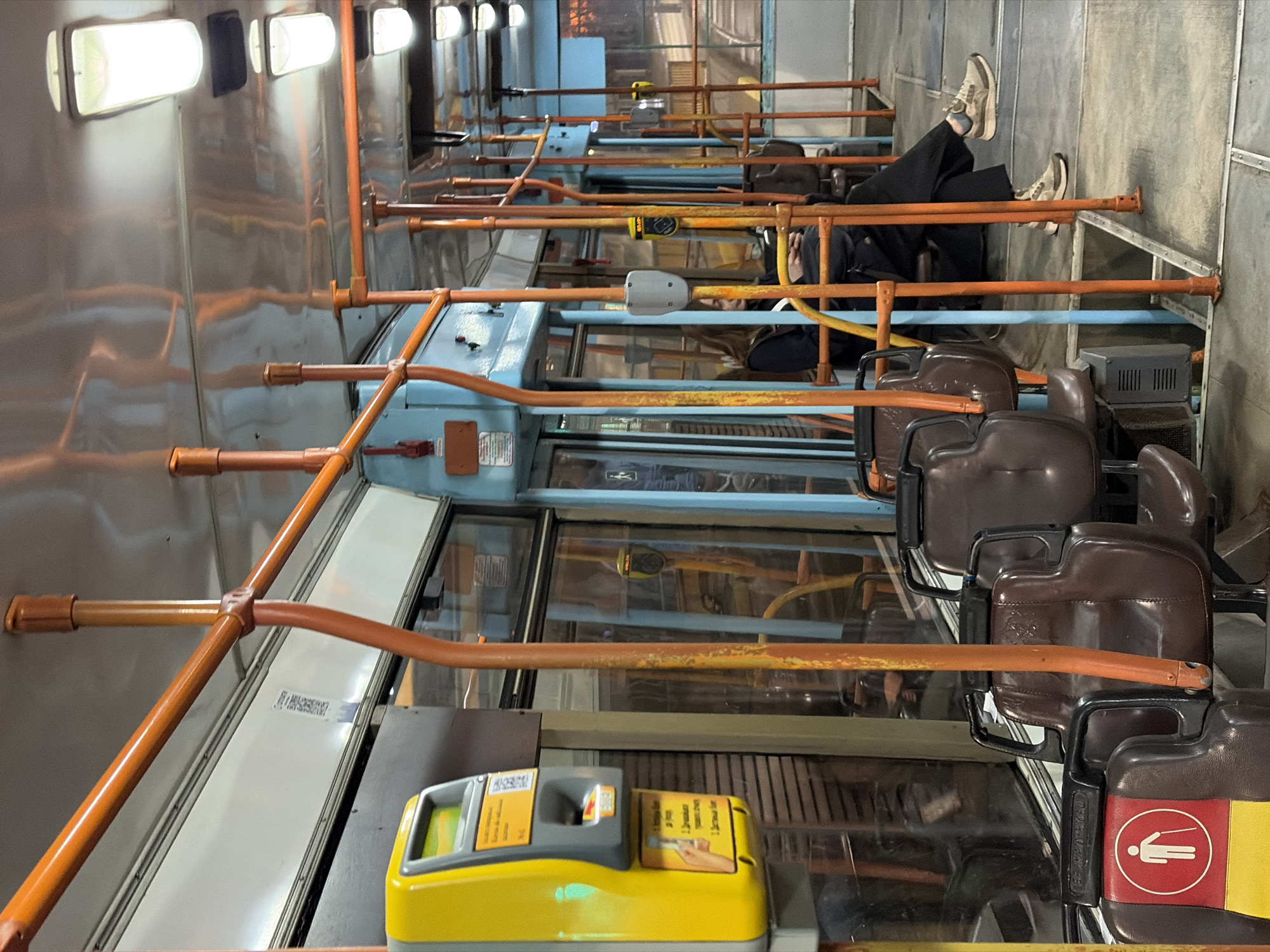 The image depicts the interior of a public transportation vehicle, likely a bus or tram, in Unknown, Unknown. The vehicle appears to be relatively empty, with rows of brown seats visible along the aisle. Orange handrails extend across the width of the vehicle, providing support for standing passengers. A yellow ticket validation machine is affixed to one of the seat structures. One person is observed reclining across multiple seats in the back of the vehicle. The vehicle's interior walls are metallic, and the windows have a blue tint. Overhead lighting fixtures are visible along the vehicle's ceiling.