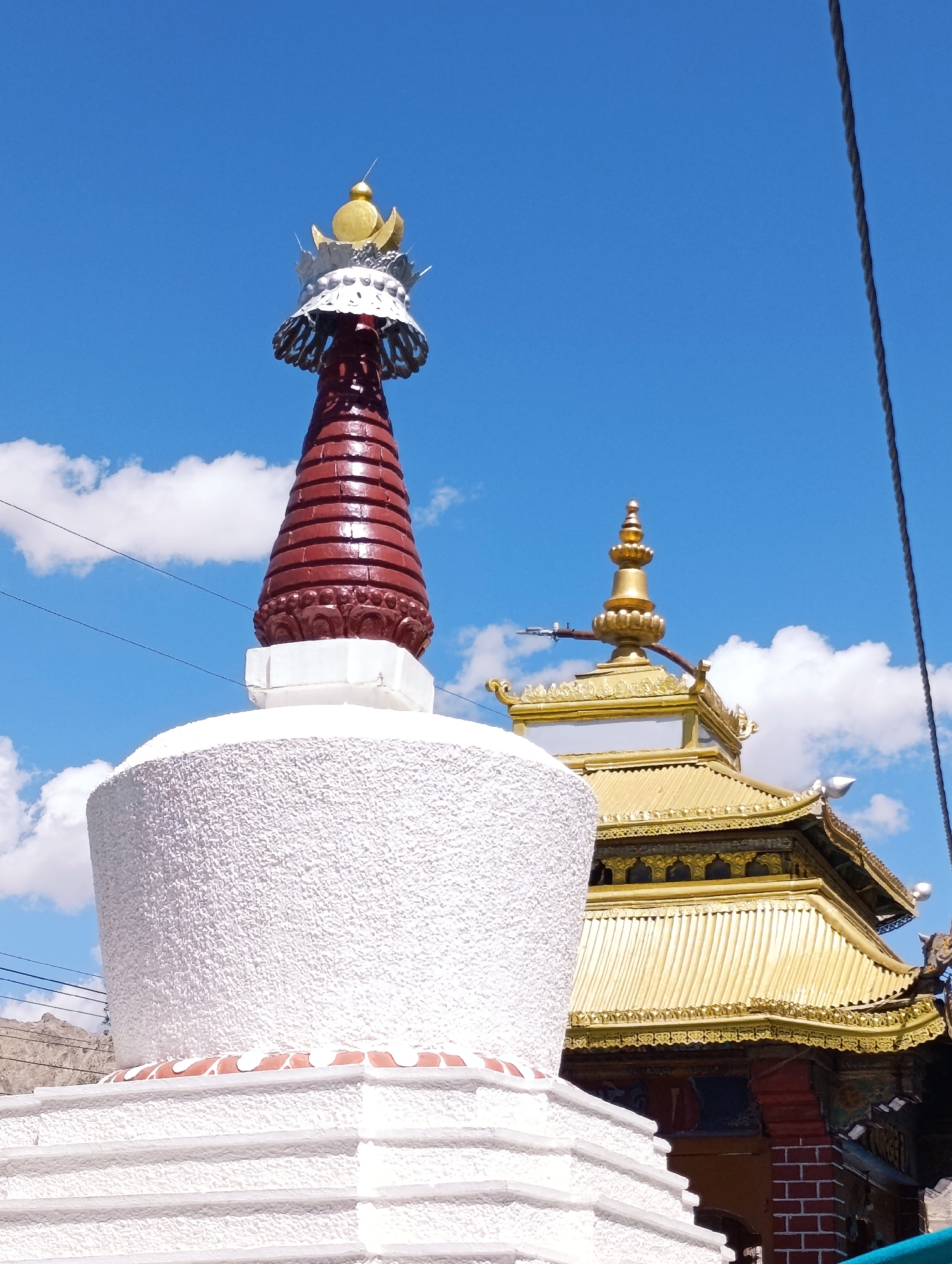 In Leh, India, a stupa stands prominently against a bright blue sky dotted with white clouds. The stupa, in the foreground, is white and textured, with distinct layered white and terracotta red bands near its base. Atop this white structure, a red conical spire rises, adorned with intricate silver filigree and a golden finial. Partially visible behind the stupa is a building with a resplendent golden roof, featuring ornate architectural details and decorative elements. The scene is partially obscured by a dark, coiled electrical wire running diagonally across the sky on the right side of the frame. In the distance, the arid, mountainous landscape of Leh is faintly visible.