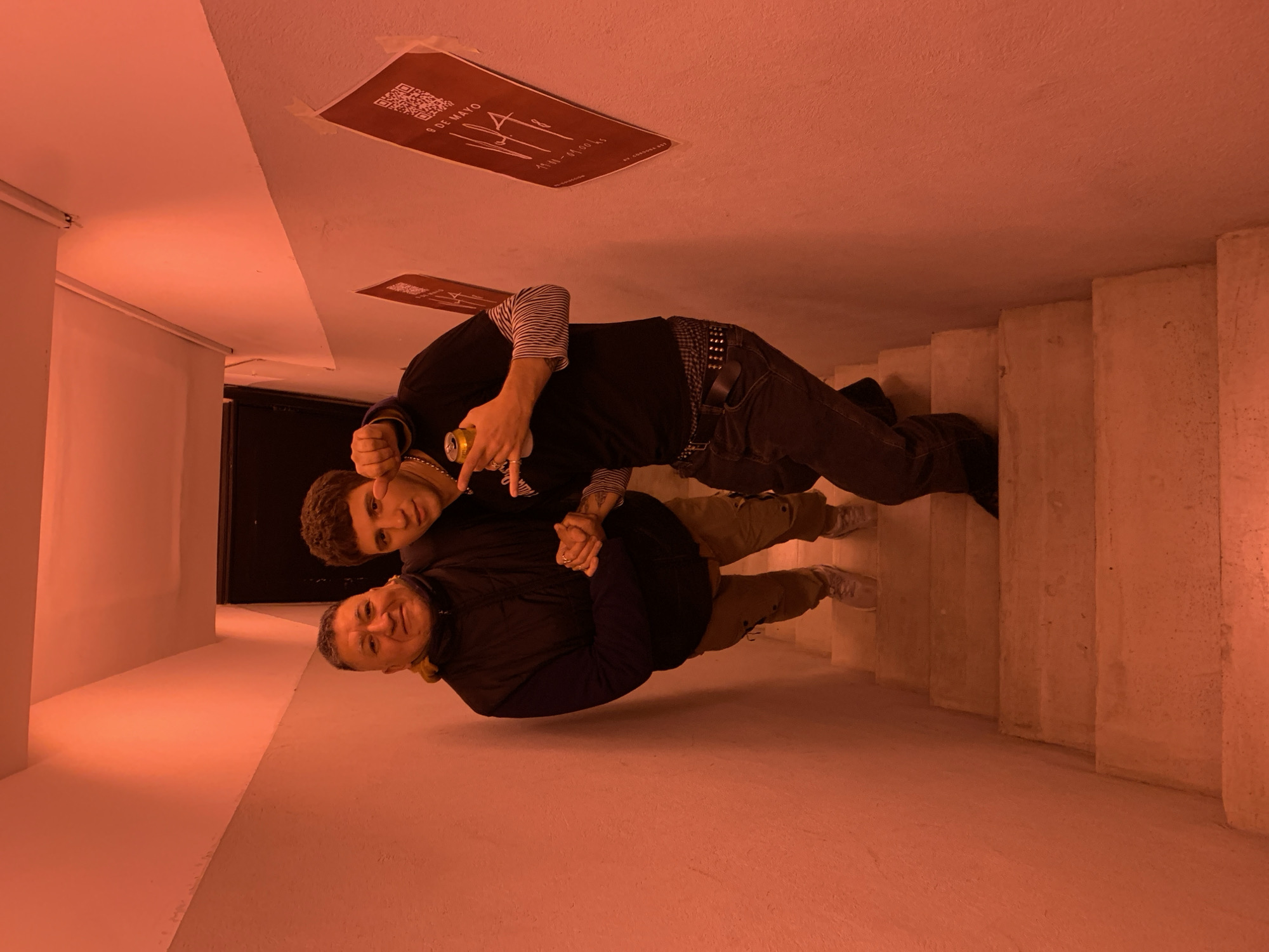 Two men are standing in a hallway and stairway bathed in red light. One man, older, is wearing a dark jacket and tan pants, holding the other by the arm. The other man, younger, is wearing a black shirt, jeans with a studded belt, and striped sleeves underneath. He holds a golden can in his other hand. The location is Buenos Aires, Argentina, indicated by a poster on the ceiling reading "9 de Mayo" above Spanish text and a QR code. The hallway leads to a dark doorway, and the right side of the image shows concrete stairs.