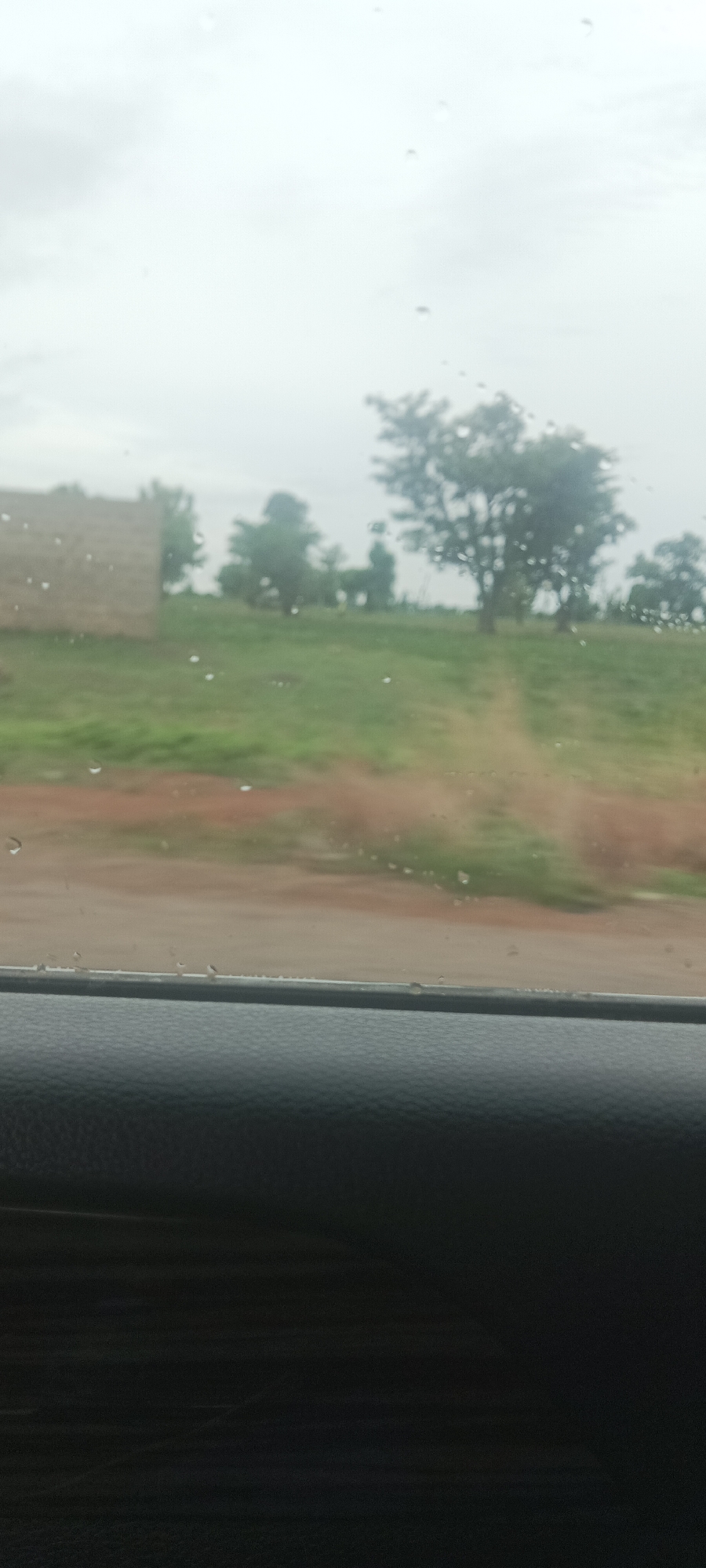 The photo was taken from inside a vehicle, likely a car, on a rainy day. Raindrops are visible on the window. The foreground consists of a dark, textured dashboard. Through the window, the scene is a rural landscape. A dirt road or path runs through the center, leading towards several trees in the distance. To the left, there is a building made of mud. The sky is overcast and grey. Based on the prompt, the location is Gwadanye, Nigeria. No people are visible in the scene.